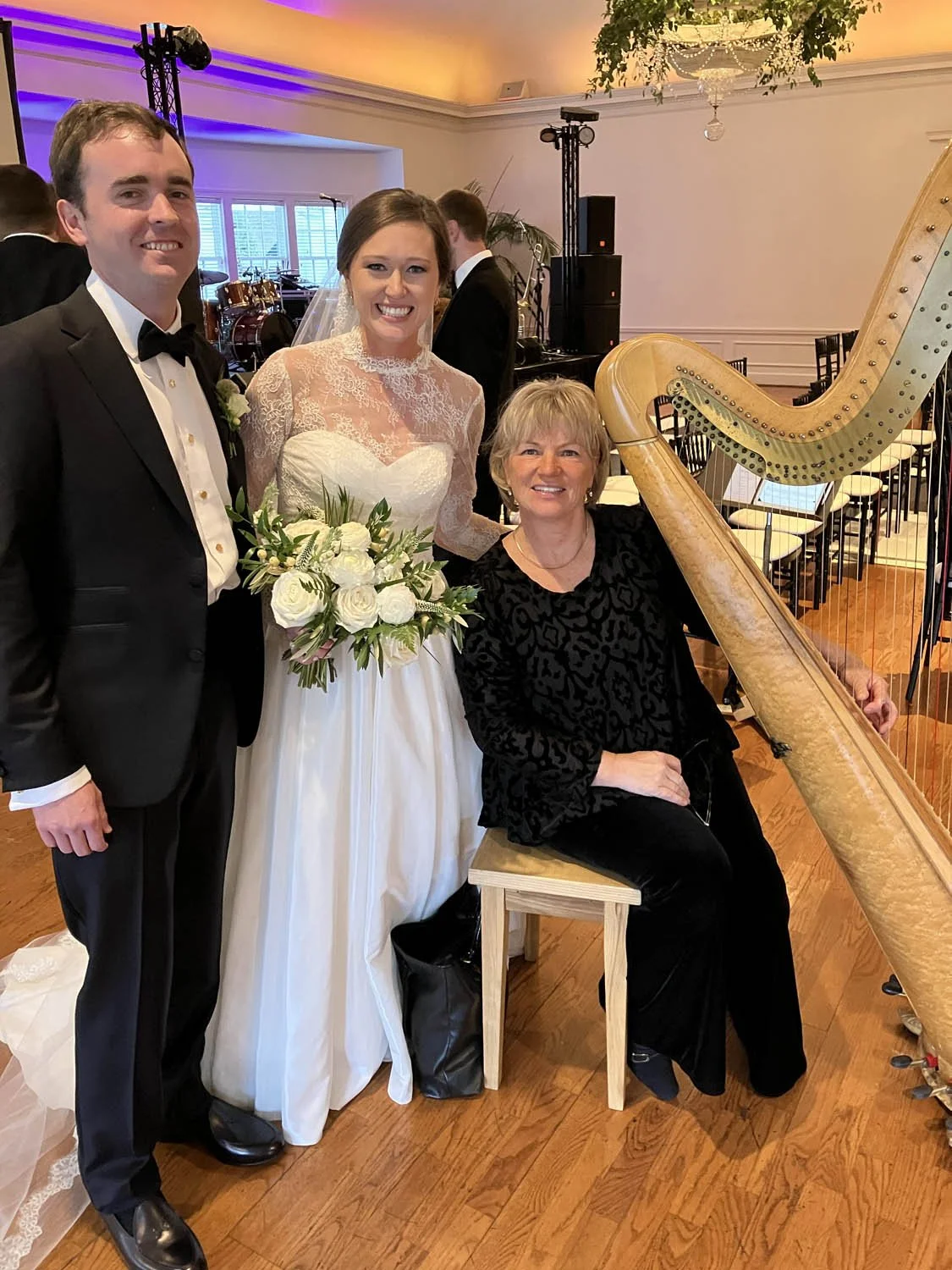 A bride and groom posing with a woman playing a harp at a wedding reception, with musical instruments and event lighting in the background.