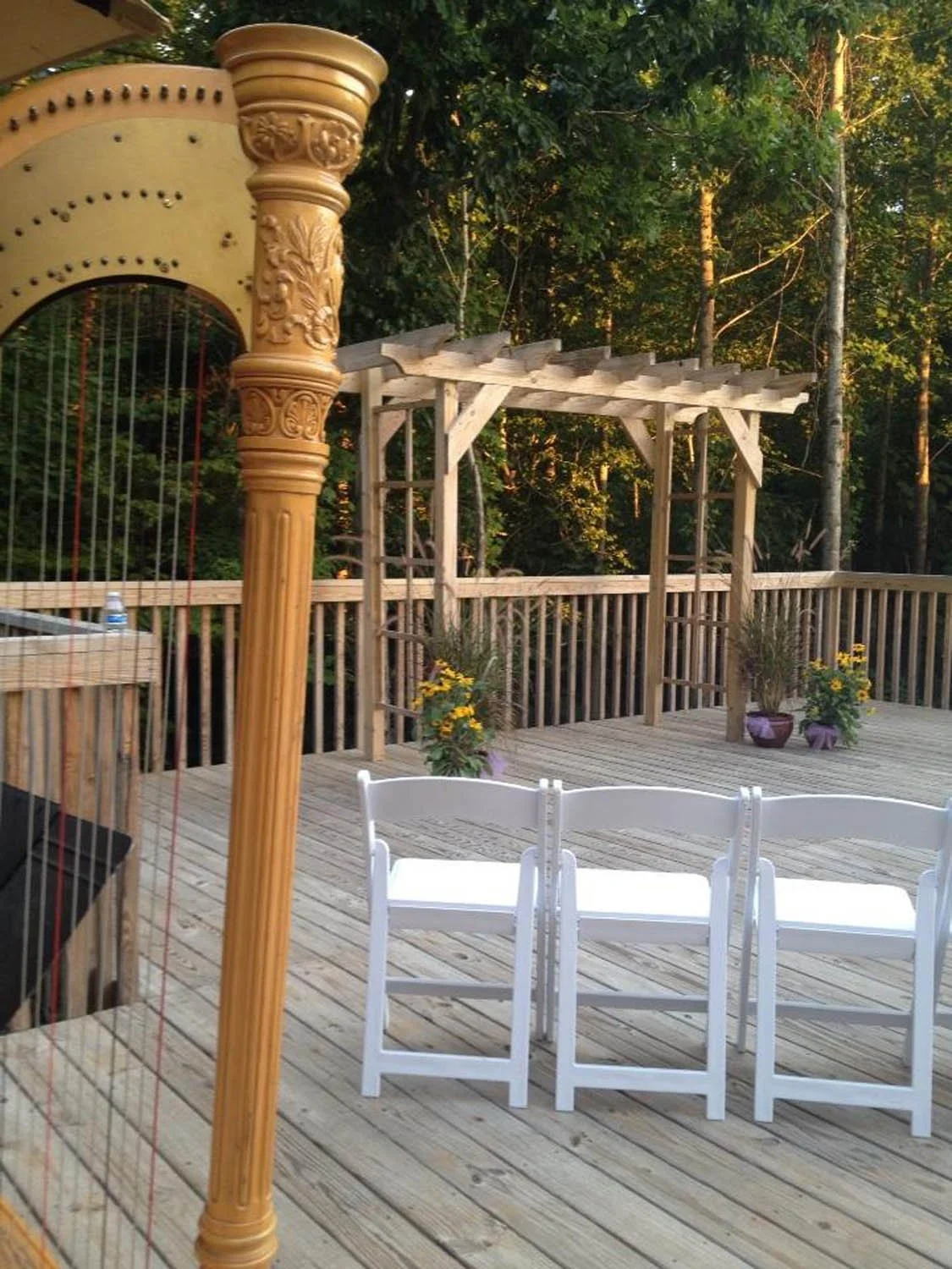 A wooden outdoor deck surrounded by trees, with a stage area, potted plants, white folding chairs, and a decorative string instrument support column.