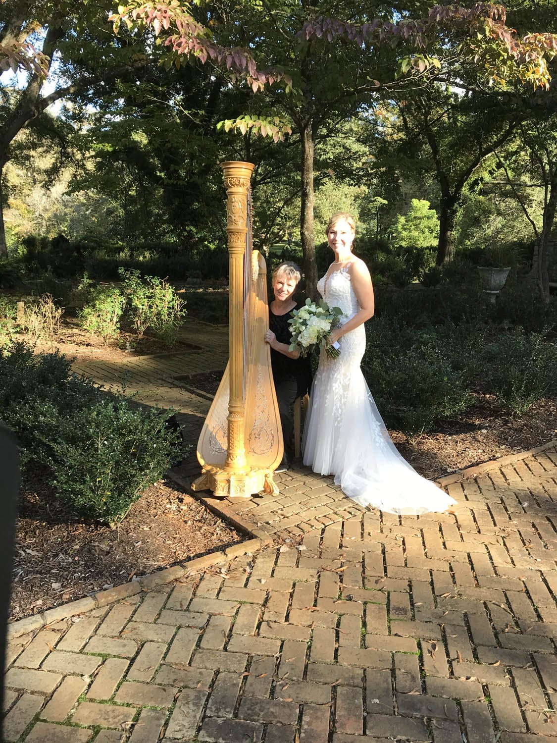 A bride in a white wedding gown holding a bouquet of white flowers standing outdoors next to a woman playing a harp, surrounded by trees and plants on a brick pathway.