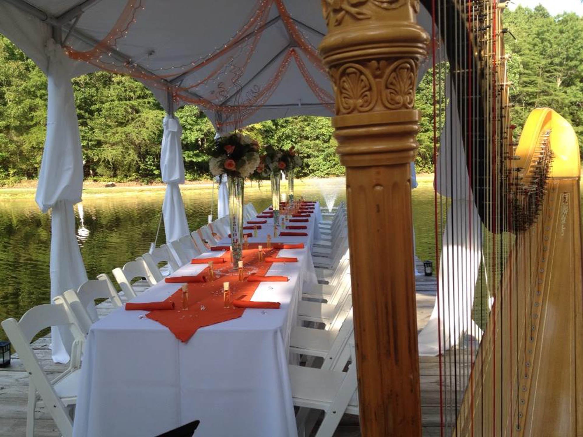 Long dinner table set up outdoors under a tent beside a lake, with white chairs, orange table runners, floral centerpieces, and a harp on the right side.