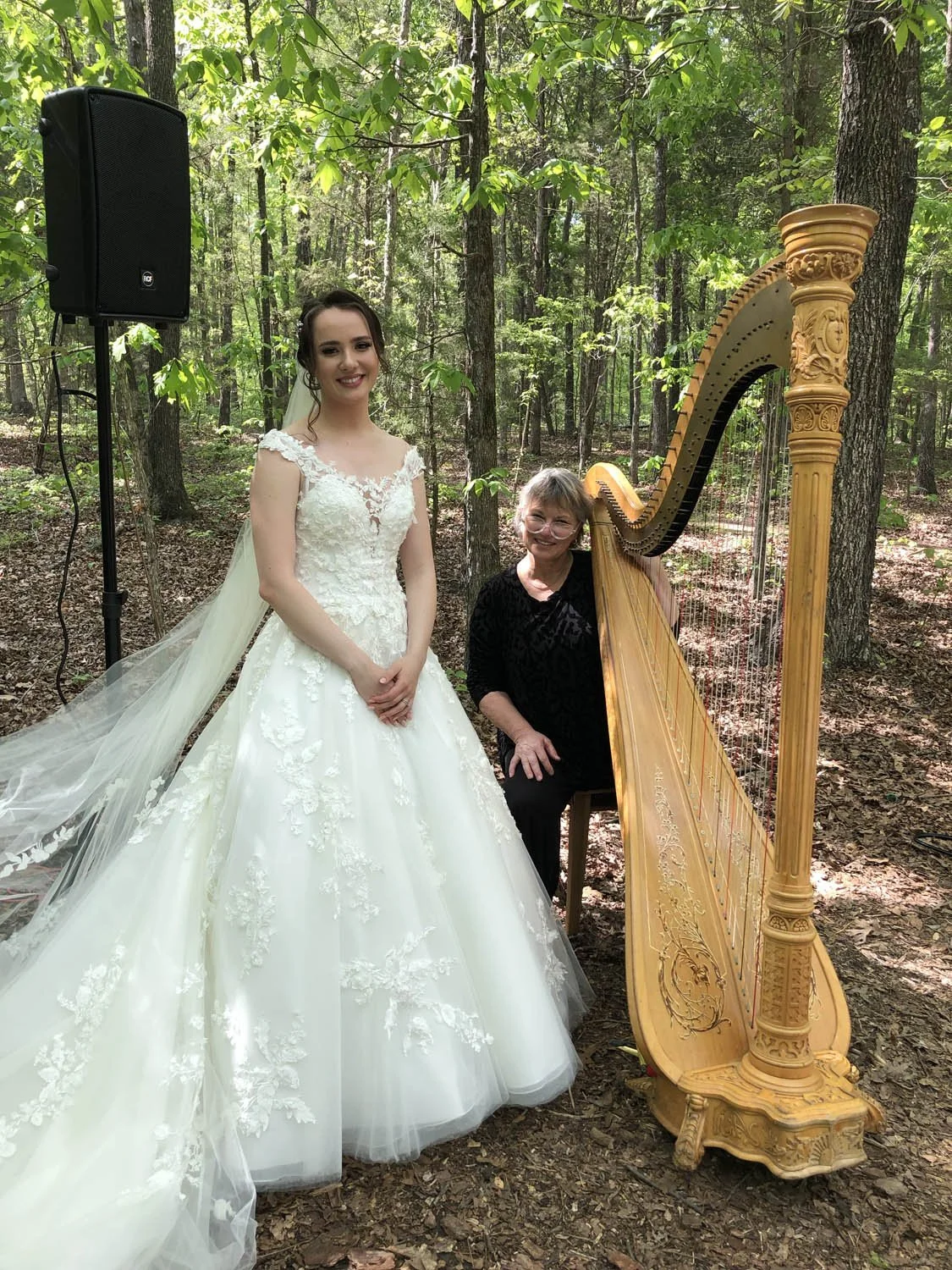 A bride in a white wedding gown with lace details standing outdoors in a forest, smiling. A woman in black seated next to a large wooden harp, also smiling. There is a black speaker on a stand behind them.