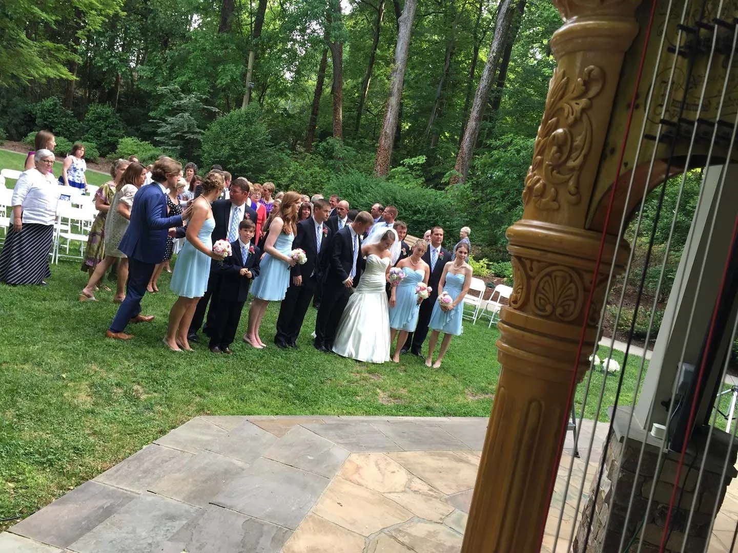 A wedding ceremony taking place outdoors in a lush green garden. The bride in a white gown, groom in a black suit, and bridesmaids in light blue dresses are standing in a line, holding flowers. Guests are seated and standing nearby, watching the ceremony.
