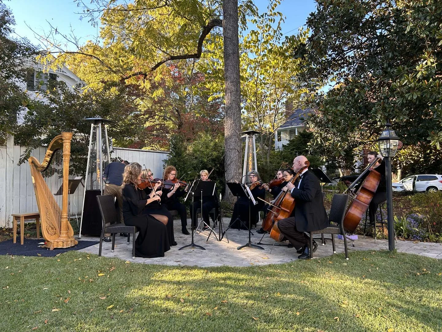 An outdoor classical music concert in a backyard featuring a string ensemble with violinists, cellists, and a harpist, set against trees and a garden with a white fence and houses in the background.