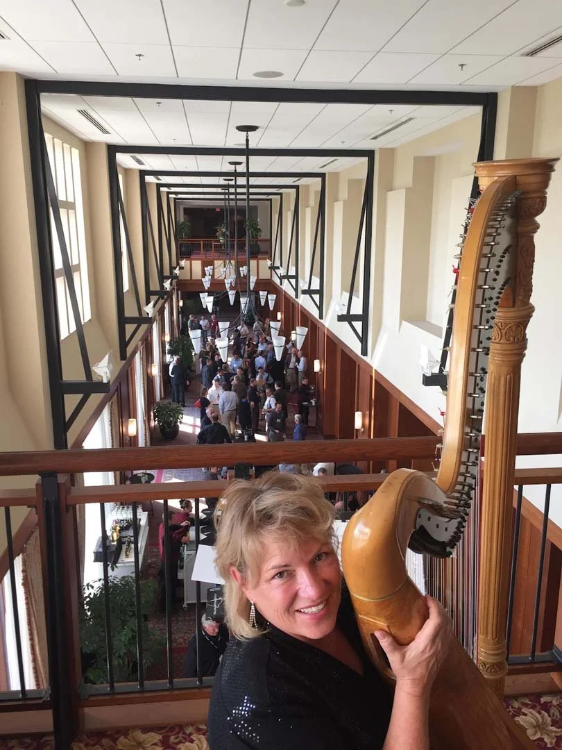 A woman smiling and holding a large wooden harp at an upper balcony overlooking a busy hotel lobby or event space with many people, tables, and hanging lights.