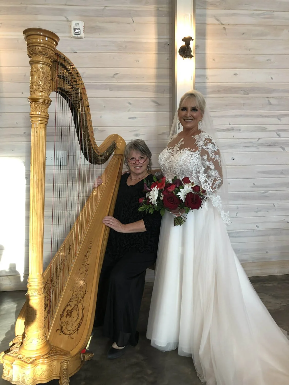 A bride in a white lace wedding dress holding a bouquet of red and white flowers, standing next to a woman in black sitting behind an ornate wooden harp.