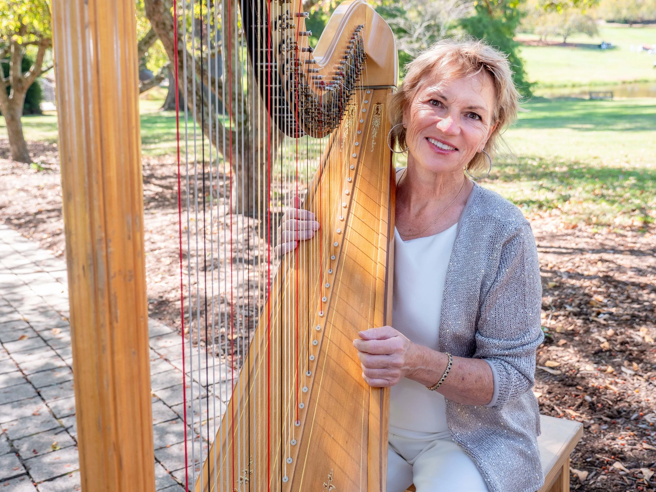 A smiling woman with short blonde hair sitting outdoors in a park, holding a wooden harp.