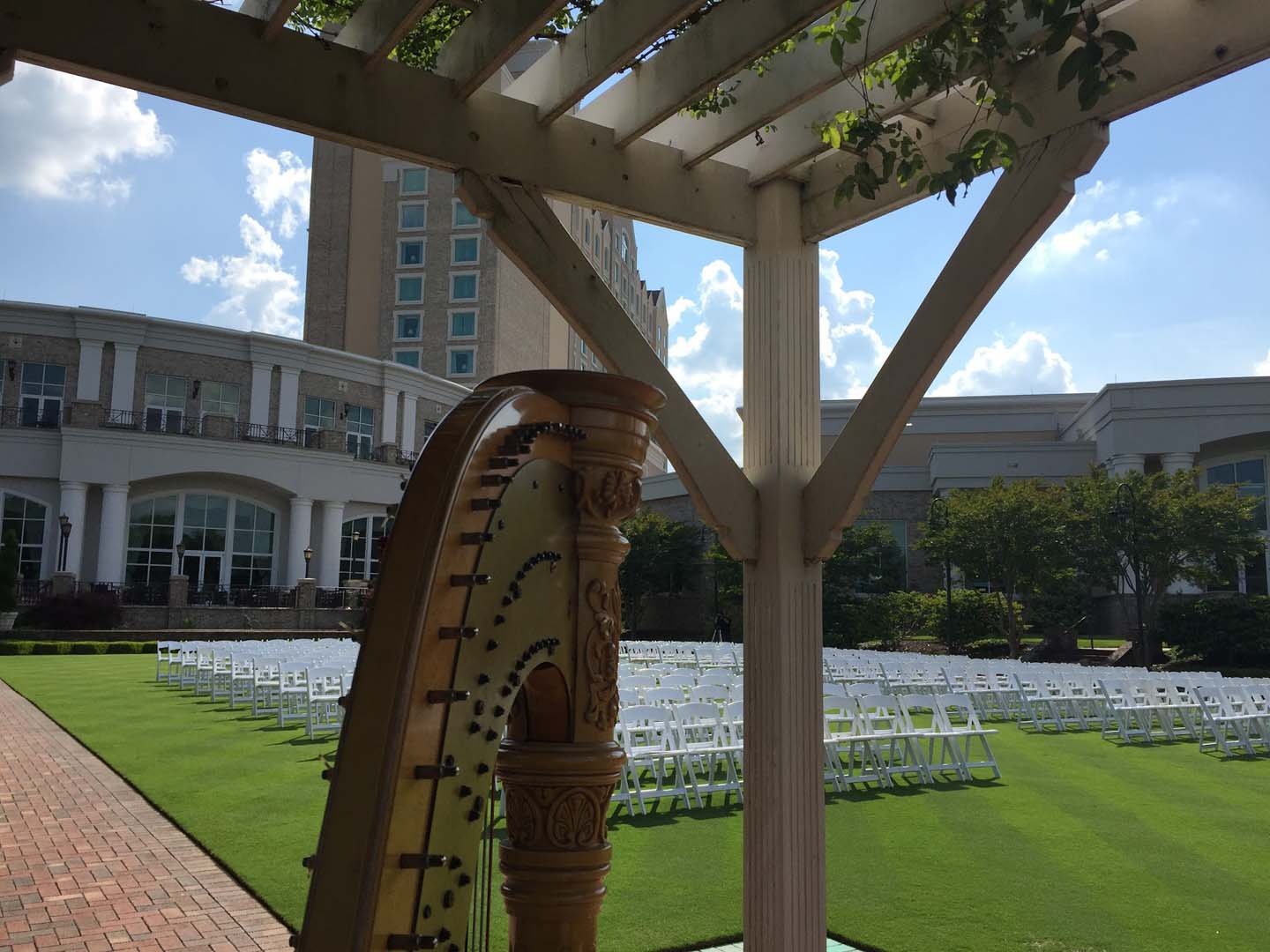 View of an outdoor event space with rows of white chairs on green lawn, a classical wooden outdoor fountain, and a building in the background under a partly cloudy sky.