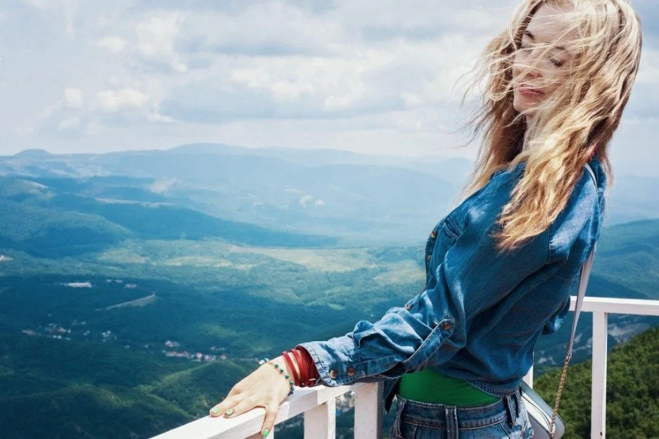 woman in blue shirt standing on a white balcony with mountains and hills in the distance