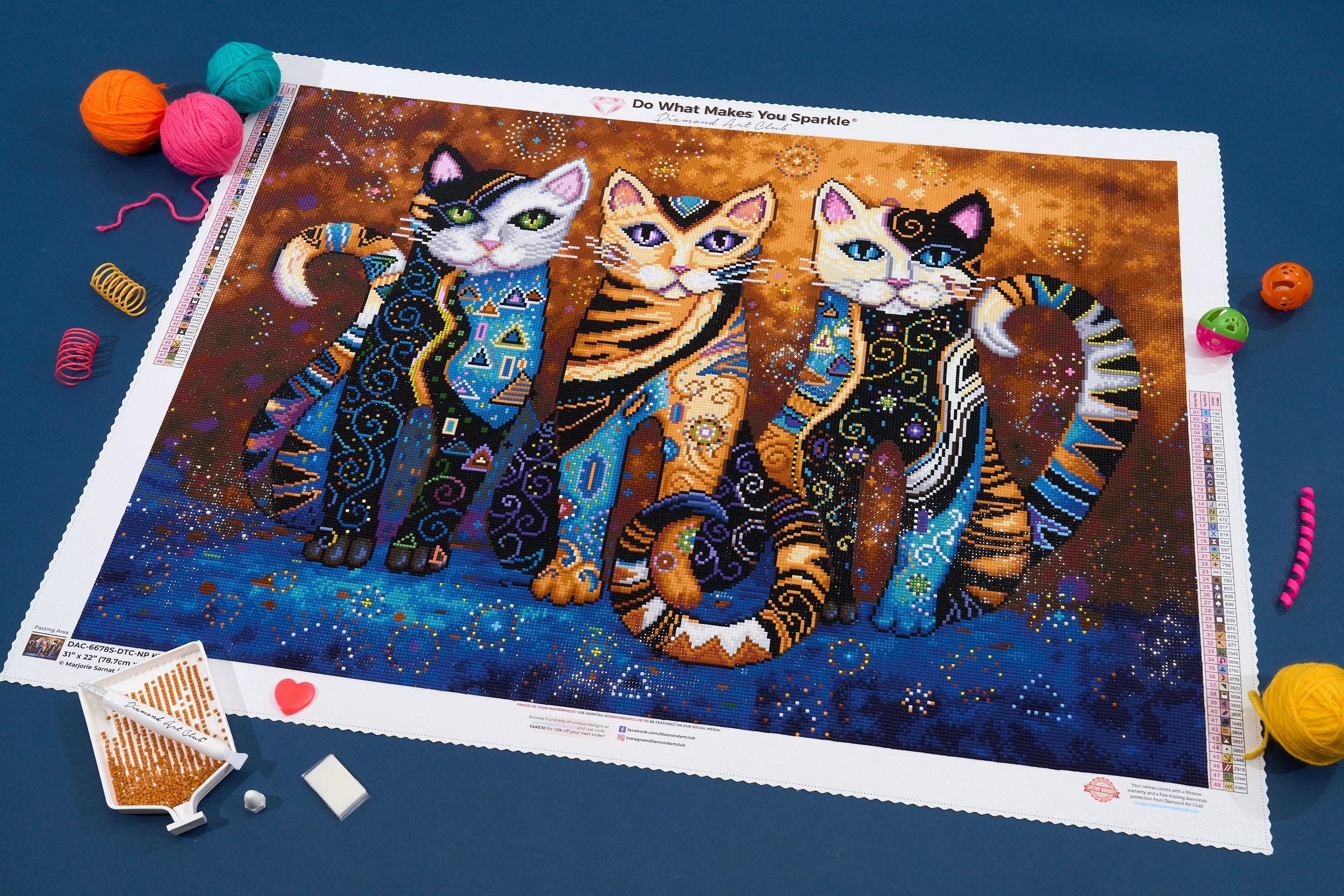 Colorful diamond paint-by-numbers artwork of four stylized cats with intricate patterns and cosmic background, surrounded by balls of yarn and small crafting accessories.