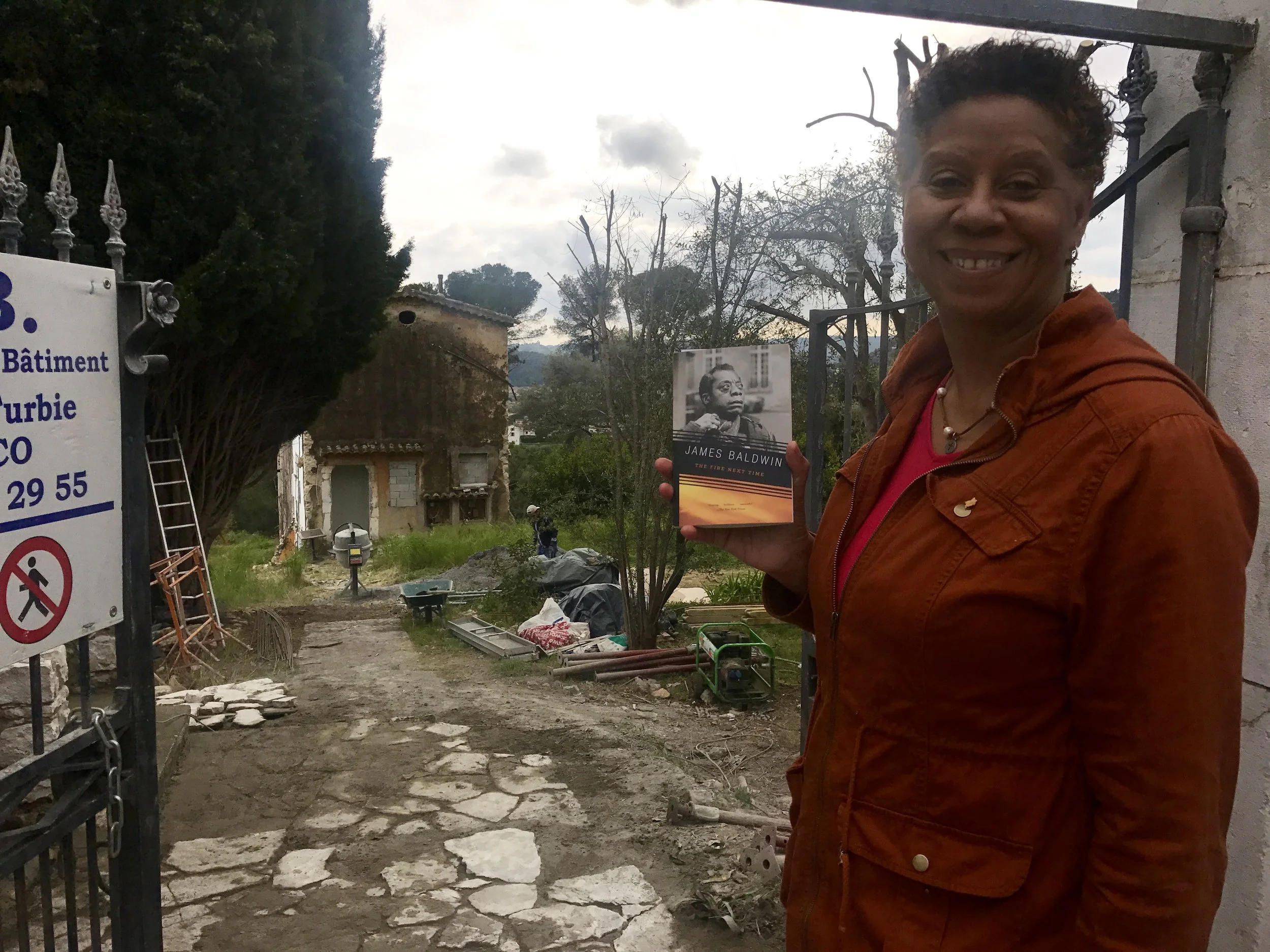 my mom holding her copy of "The Fire Next Time" by James Baldwin in front of his home.