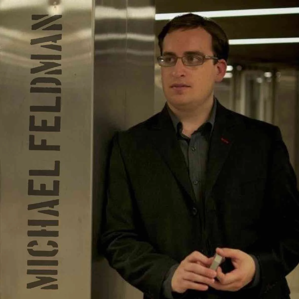 Michael Feldman Headshot holding cards leaning against a metal column