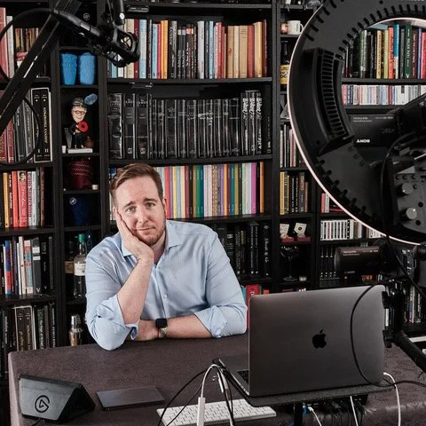 James Alan sitting in his home studio with laptop and ring light in front of large bookcases