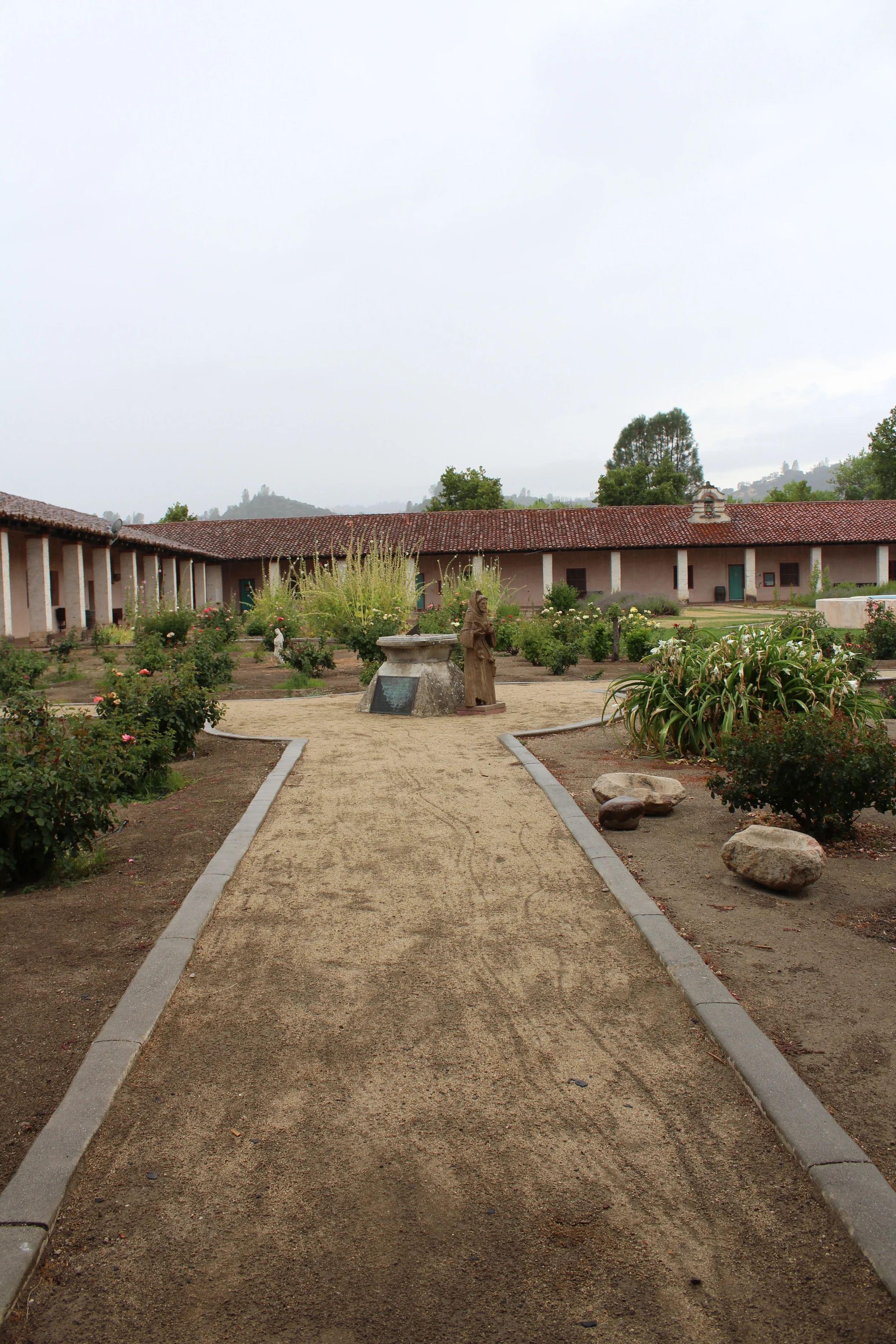 This photo was taken from the steps of the church , facing into the Padres' Garden. You are looking at a sundial and a wooden statue of St. Anthony, the saint that this mission was named for. The courtyard is planted with all colors of roses, and have been lovingly maintained by a local lady in her 80s. Every year toward the end of January the mission sells cuttings of its roses as an ongoing fund raiser for the mission's seismic retrofit. In the distance you can see the Santa Lucia coast range, very much covered by the rain clouds. 