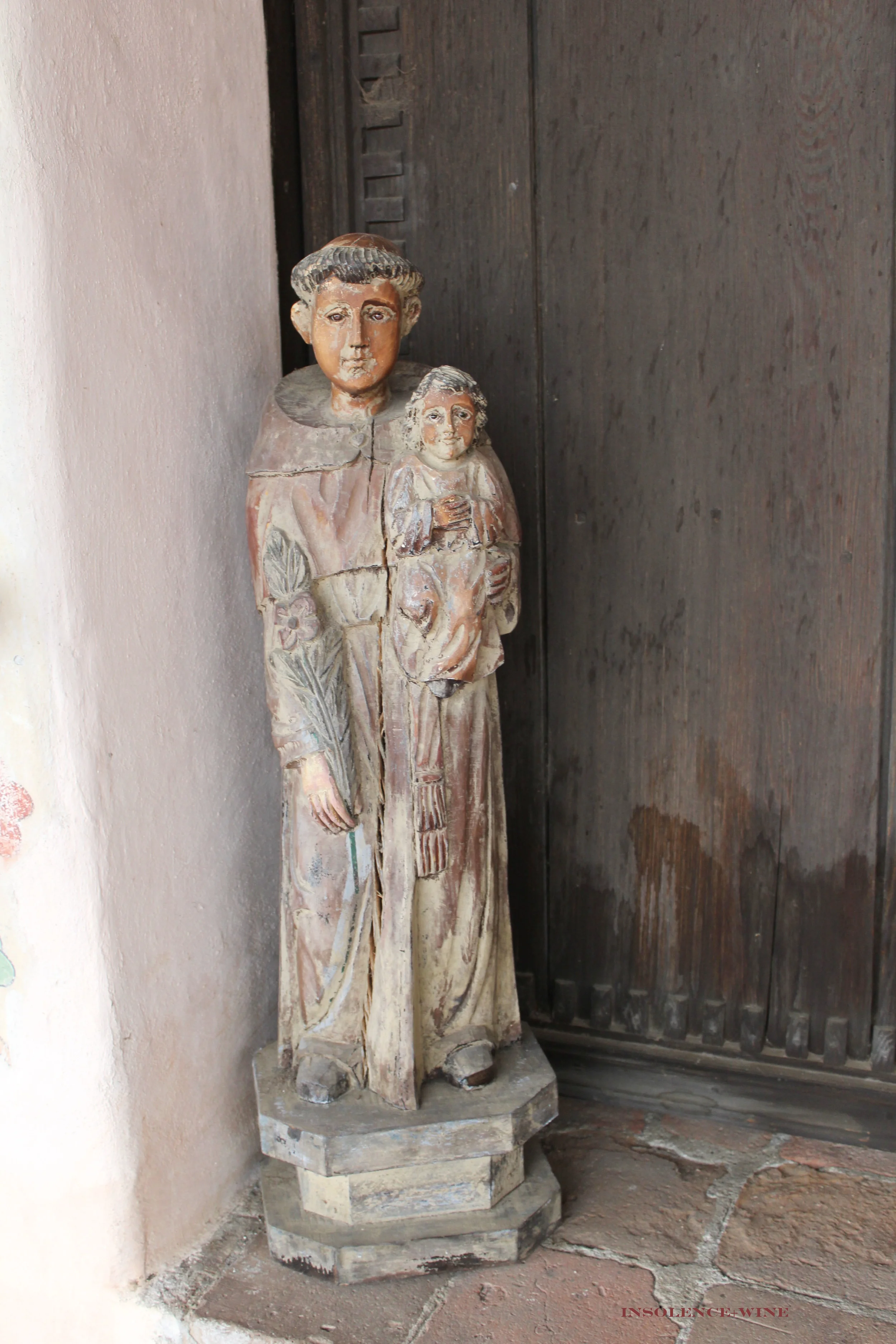 Wooden statuette of St. Anthony de Padua on the steps outside of the church, facing into the courtyard. 