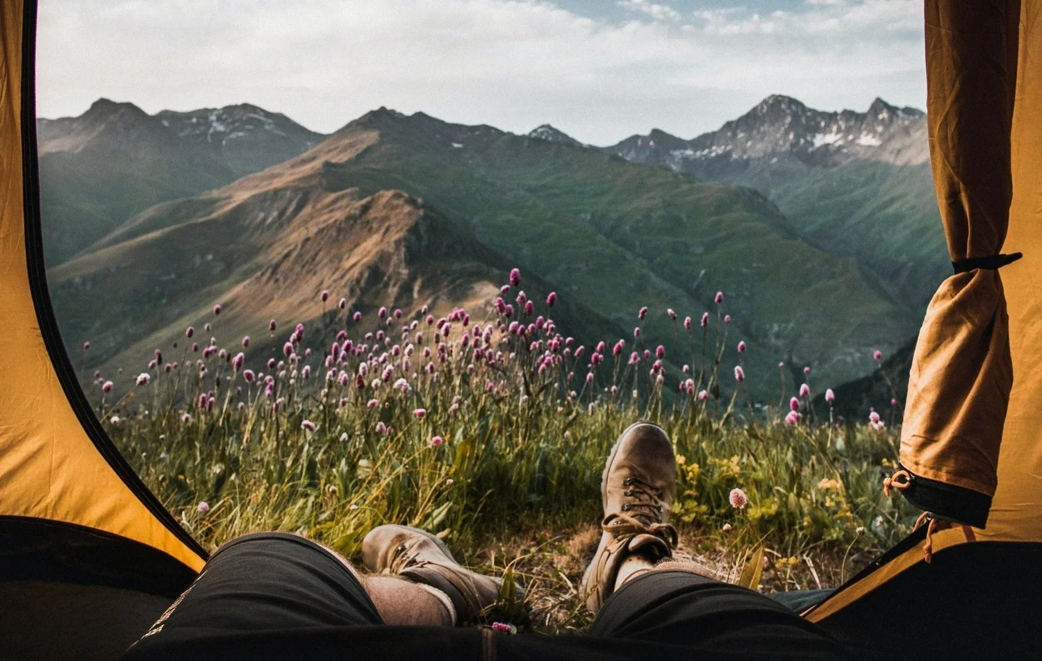 View from inside a tent of a person’s legs and hiking boots, overlooking a mountainous landscape with green hills and purple wildflowers under a cloudy sky.