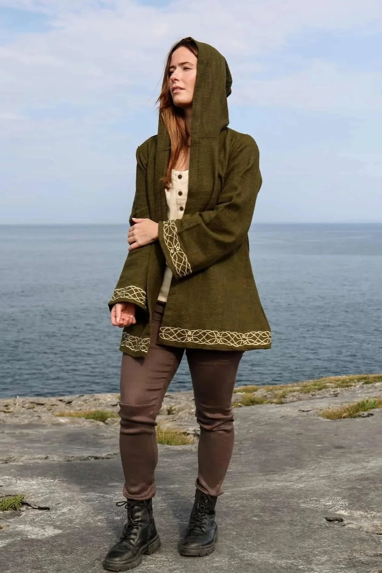 Woman wearing Celtic Fusions handwoven cotton hooded wrap cardigan with Celtic knotwork embroidery on cuffs and hem; belted at waist. Folklore cardigan photographed in a woodland setting.
