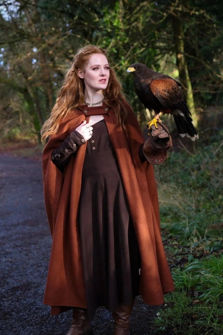 Woman wearing a hooded cape styled as a medieval cloak by Celtic Fusion, evoking a red riding hood silhouette in a rustic stone ruin setting