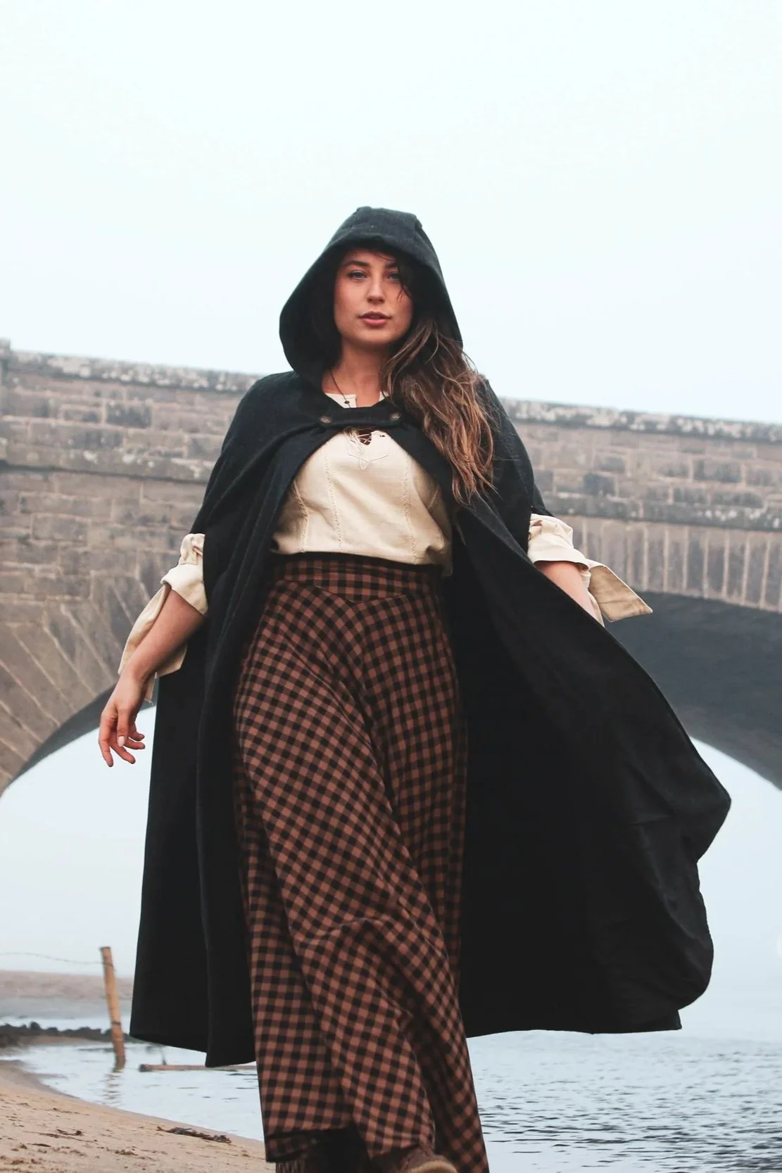 Woman wearing a hooded cape styled as a medieval cloak by Celtic Fusion, evoking a red riding hood silhouette in a rustic stone ruin setting