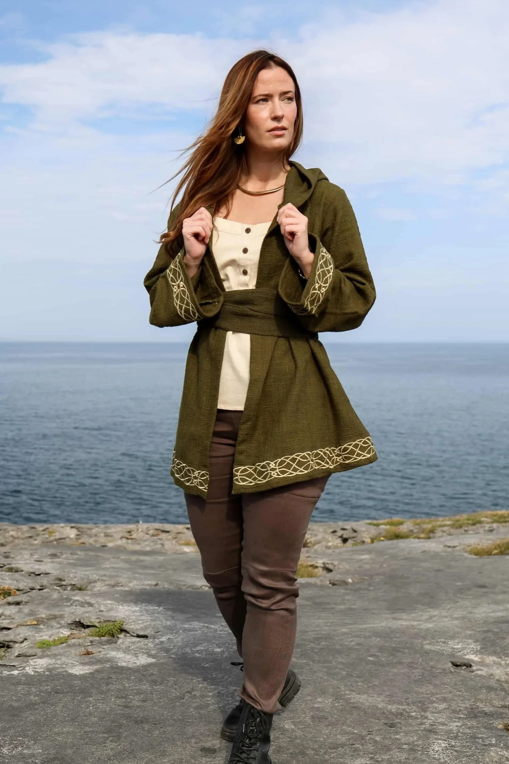 Woman wearing Celtic Fusions handwoven cotton hooded wrap cardigan with Celtic knotwork embroidery on cuffs and hem; belted at waist. Folklore cardigan photographed in a woodland setting.