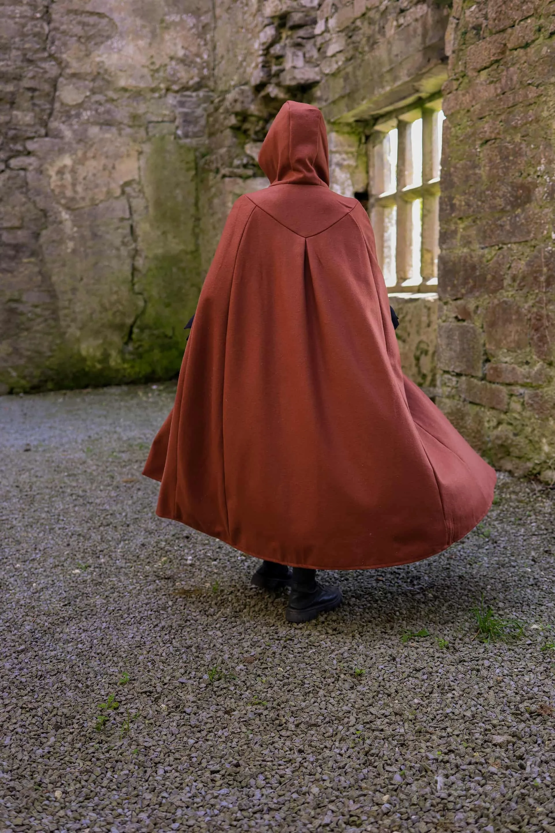 Woman wearing a hooded cape styled as a medieval cloak by Celtic Fusion, evoking a red riding hood silhouette in a rustic stone ruin setting