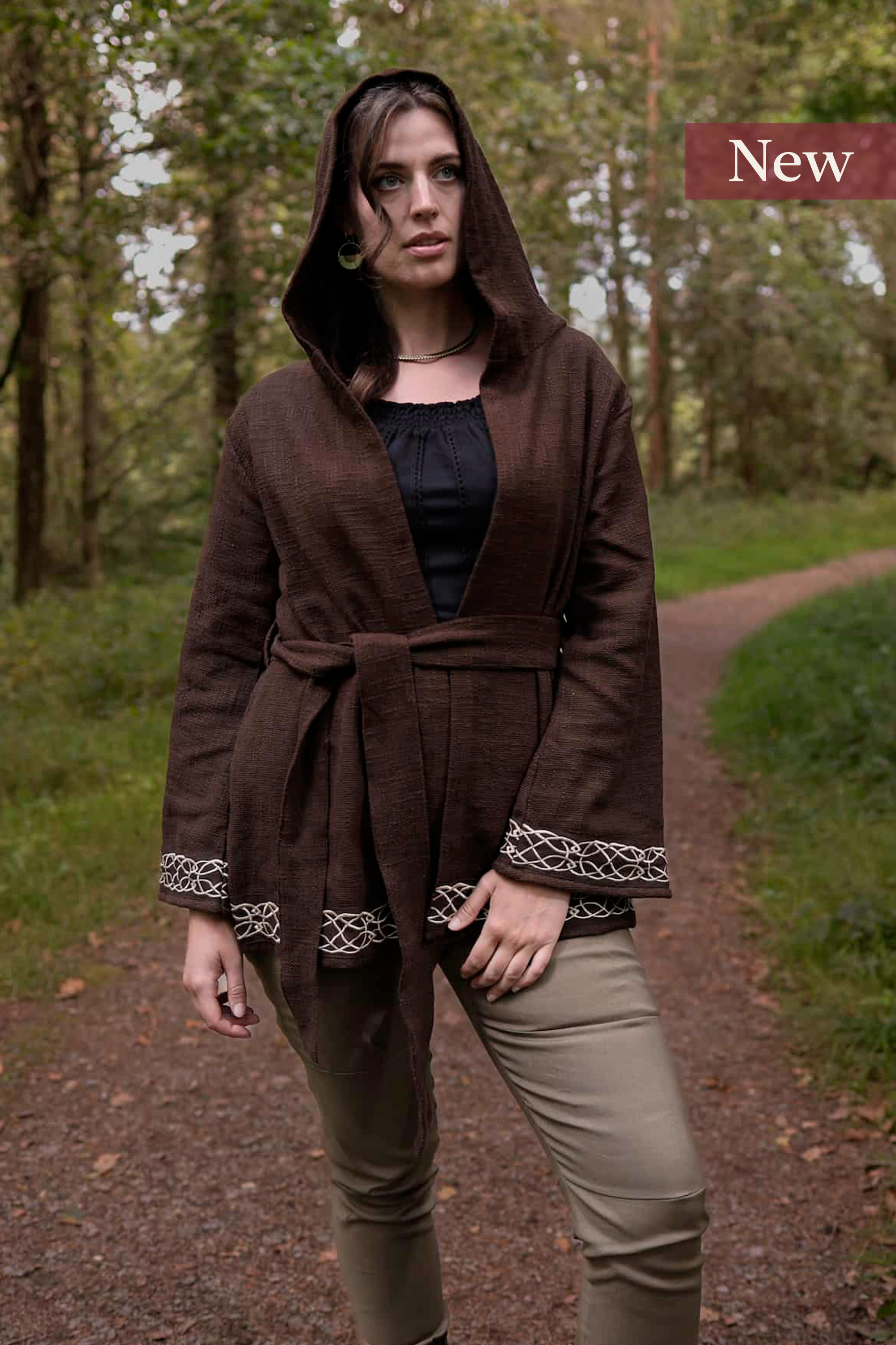 Woman wearing Celtic clothing handwoven wrap cardigan with Celtic knot embroidery on cuffs and hem; belted at waist. Folklore cardigan photographed in a woodland setting.