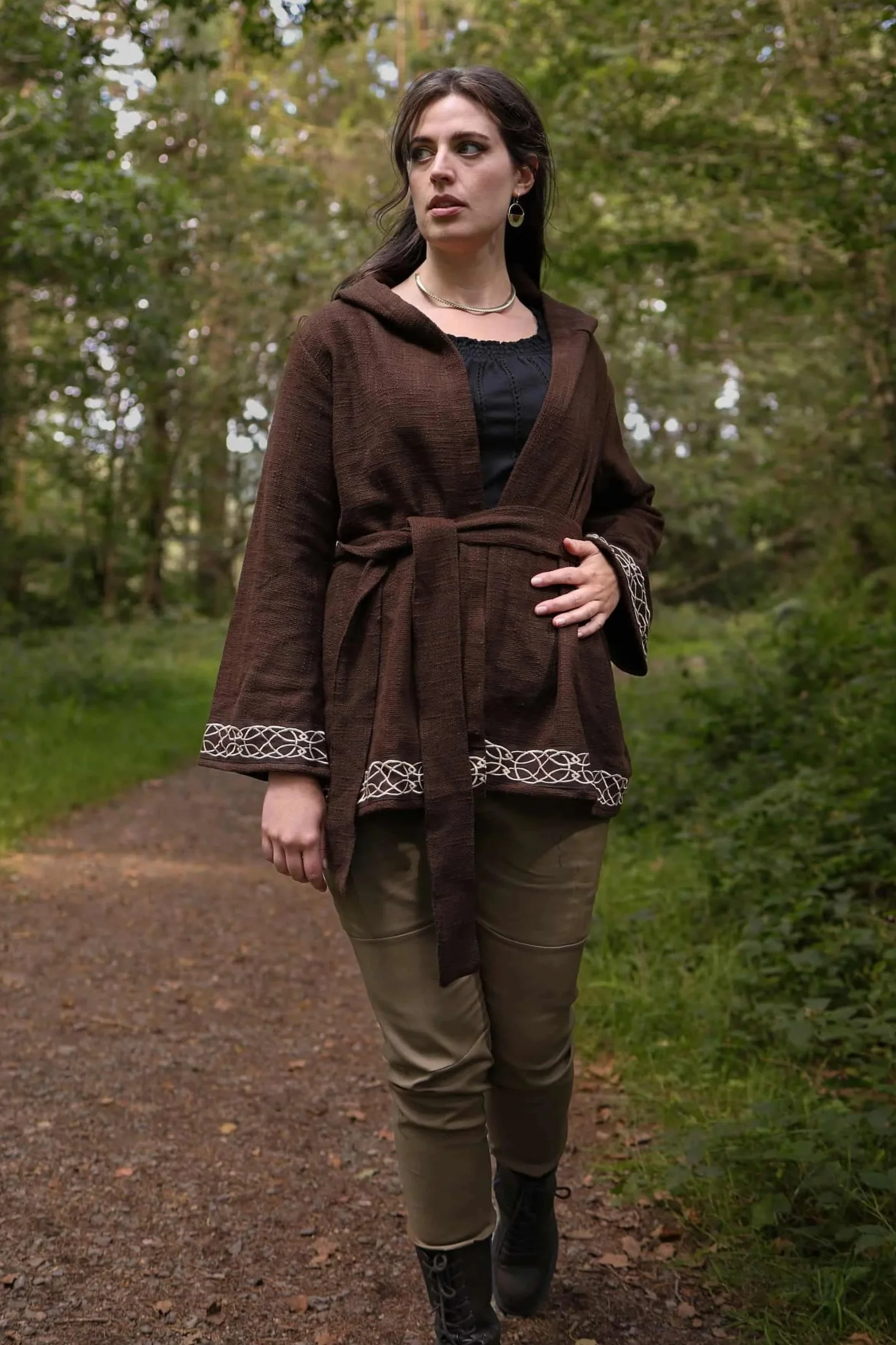Woman wearing Celtic clothing handwoven wrap cardigan with Celtic knot embroidery on cuffs and hem; belted at waist. Folklore cardigan photographed in a woodland setting.