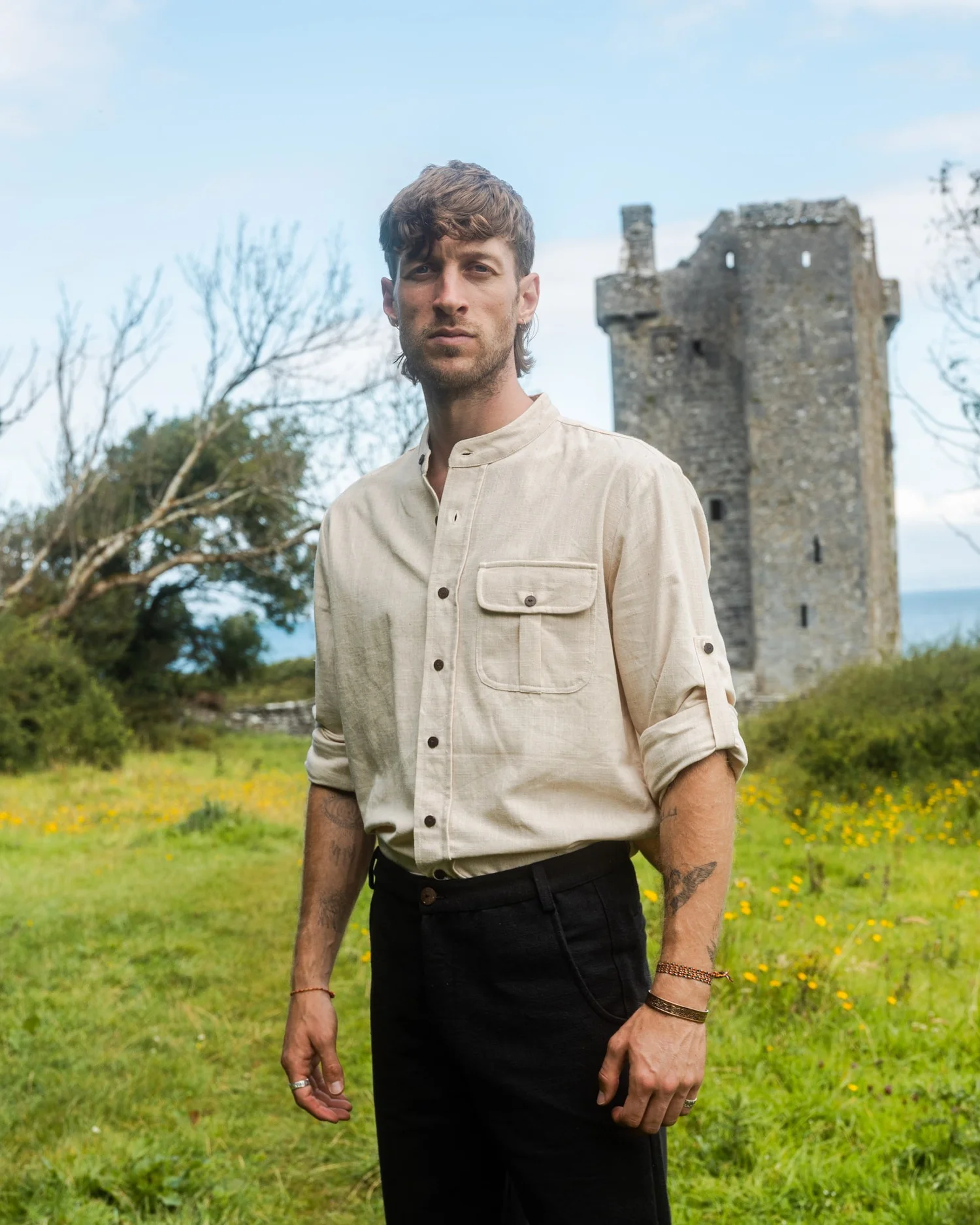 Cream collarless shirt with dark trousers beside an old stone tower