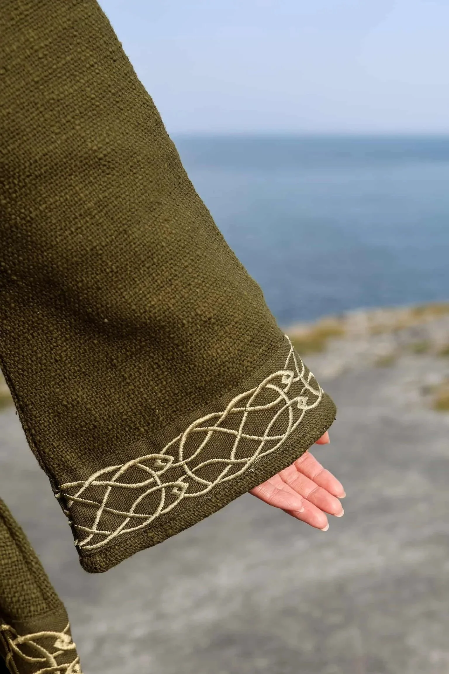 Woman wearing Celtic Fusions handwoven cotton hooded wrap cardigan with Celtic knotwork embroidery on cuffs and hem; belted at waist. Folklore cardigan photographed in a woodland setting.