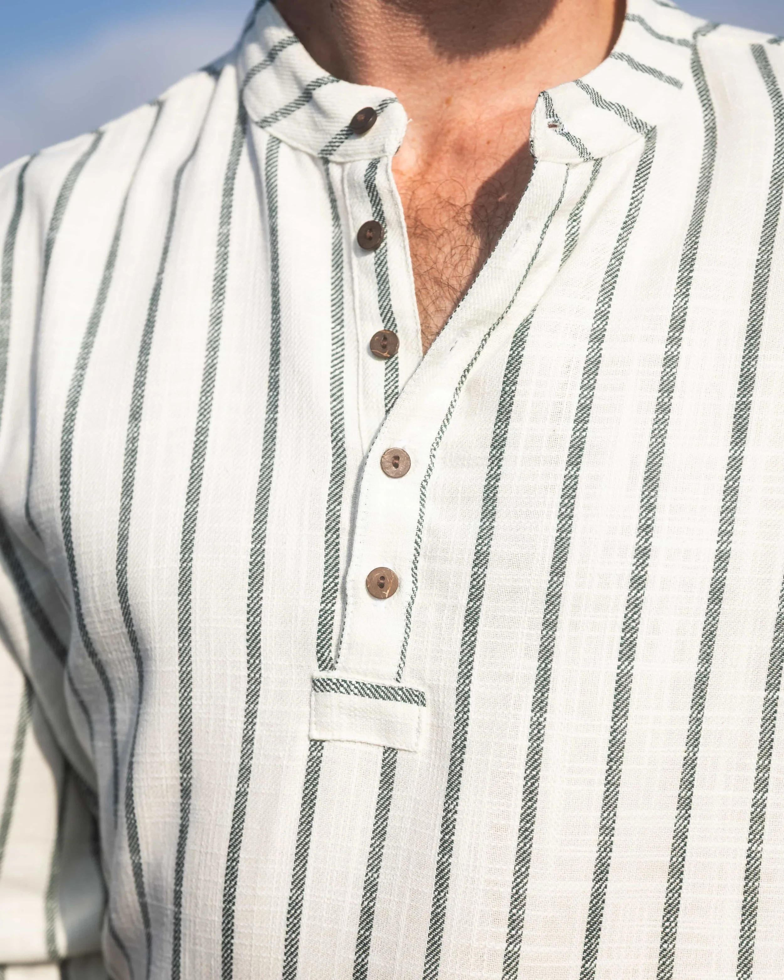 Model wearing a forest green and cream striped collarless shirt in 100% cotton, with coconut buttons and rolled sleeves, pictured by the Atlantic coast.