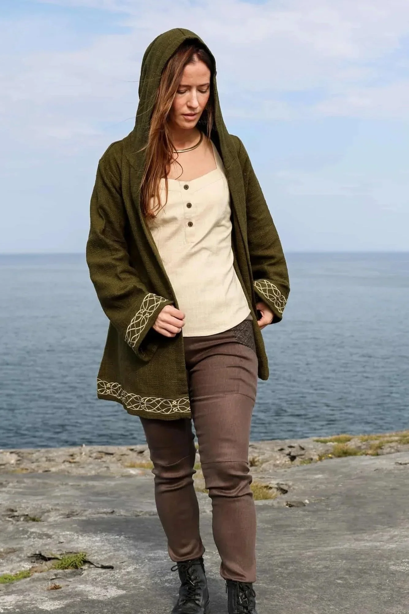 Woman wearing Celtic Fusions handwoven cotton hooded wrap cardigan with Celtic knotwork embroidery on cuffs and hem; belted at waist. Folklore cardigan photographed in a woodland setting.