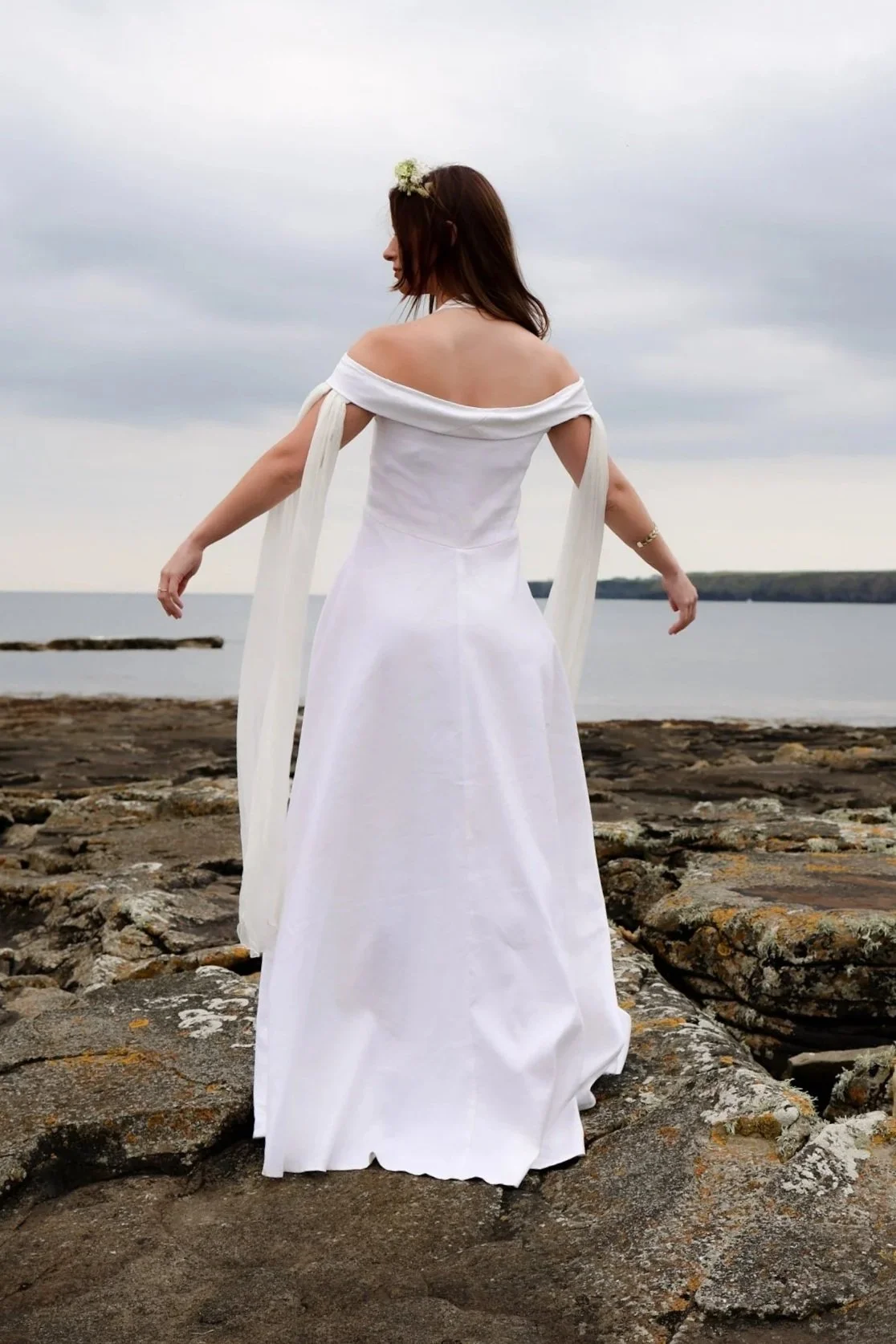 White halterneck linen wedding dress with off-shoulder wrap and flowing chiffon sleeves. Figure-hugging fit with full skirt, sustainably made and perfect for Celtic-inspired ceremonies.