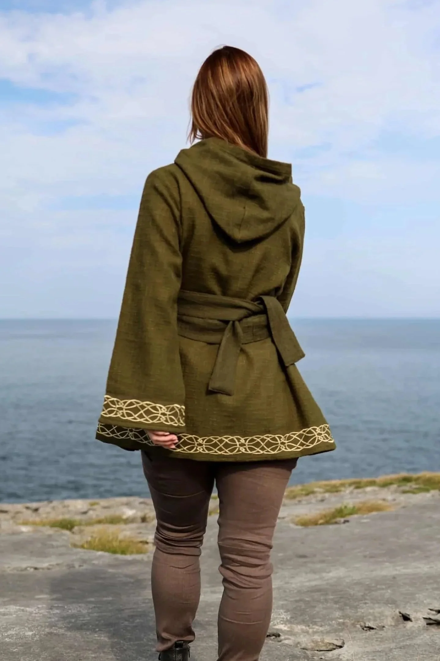 Woman wearing Celtic Fusions handwoven cotton hooded wrap cardigan with Celtic knotwork embroidery on cuffs and hem; belted at waist. Folklore cardigan photographed in a woodland setting.