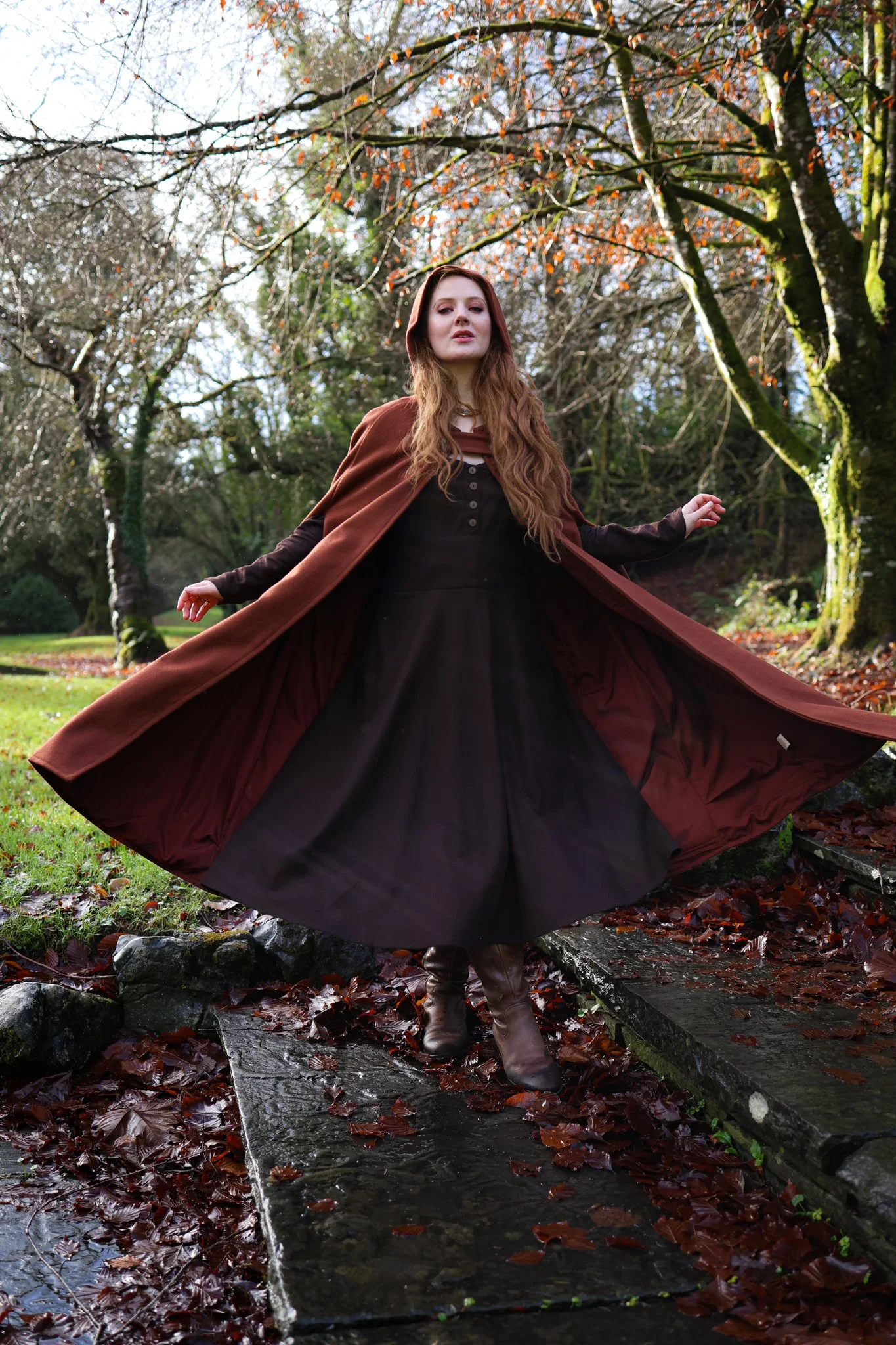 Woman wearing a hooded cape styled as a medieval cloak by Celtic Fusion, evoking a red riding hood silhouette in a rustic stone ruin setting
