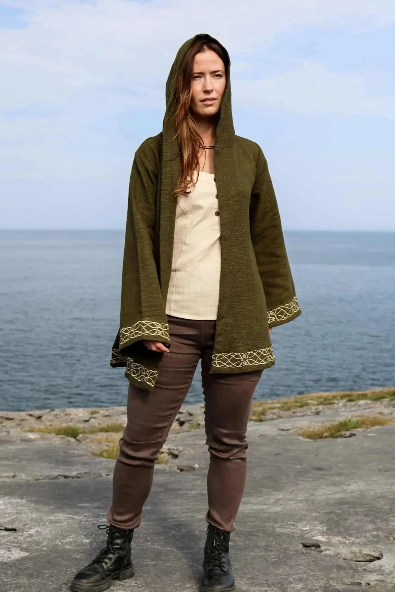 Woman wearing Celtic Fusions handwoven cotton hooded wrap cardigan with Celtic knotwork embroidery on cuffs and hem; belted at waist. Folklore cardigan photographed in a woodland setting.