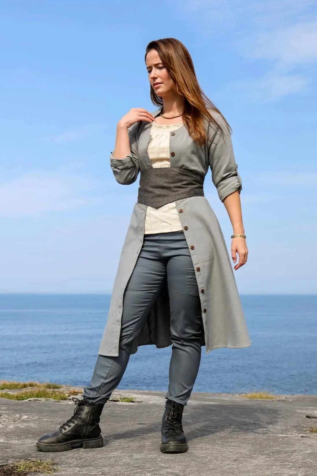 Model wearing a blue cotton button down dress with Celtic trim, styled as a midi dress with pockets and detailed button front dress closure.