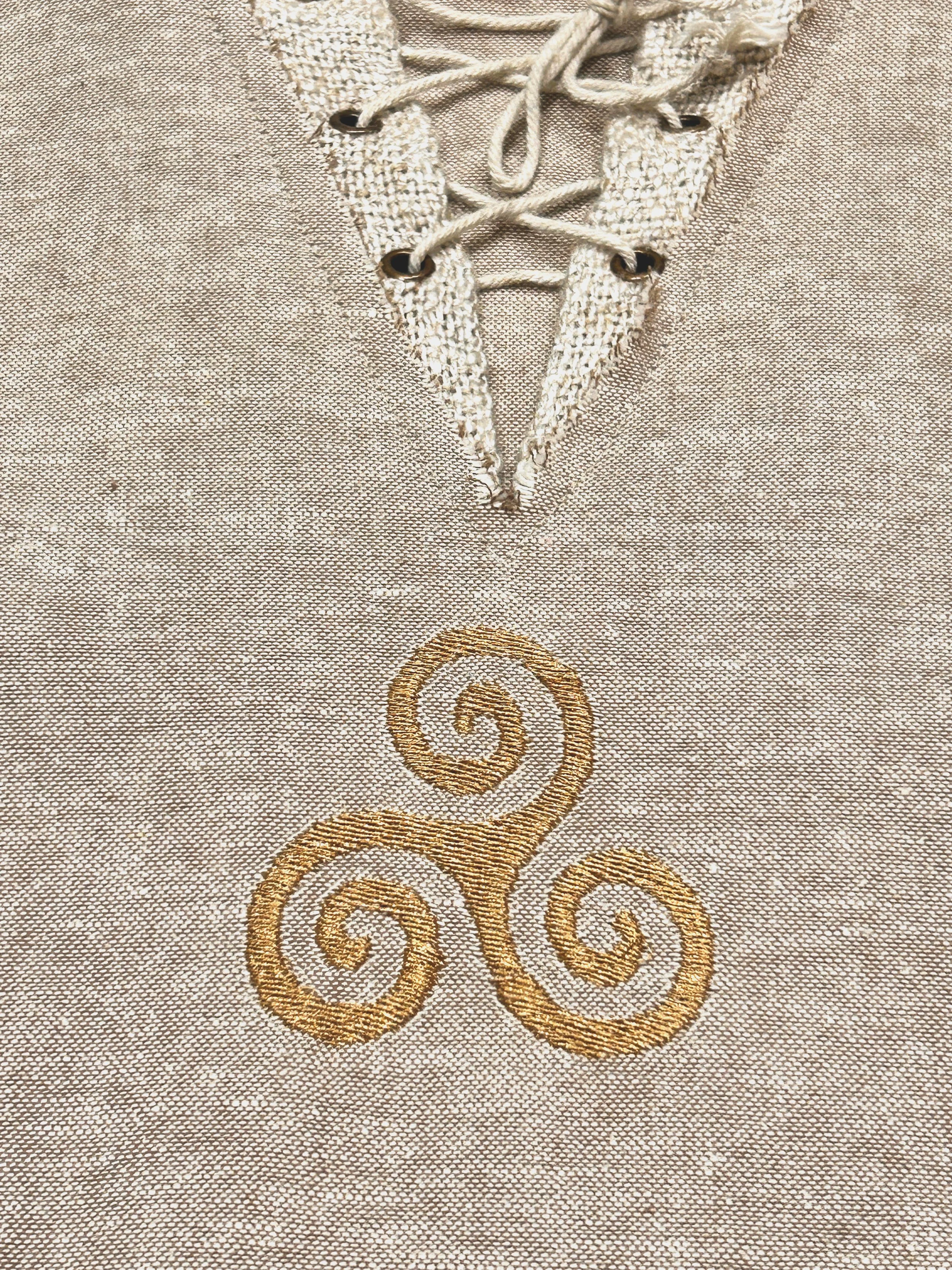 A male model wears a natural beige Celtic shirt with a lace-up neckline and golden Triskelion embroidery, styled with forest green trousers on the cliffs of Ireland.