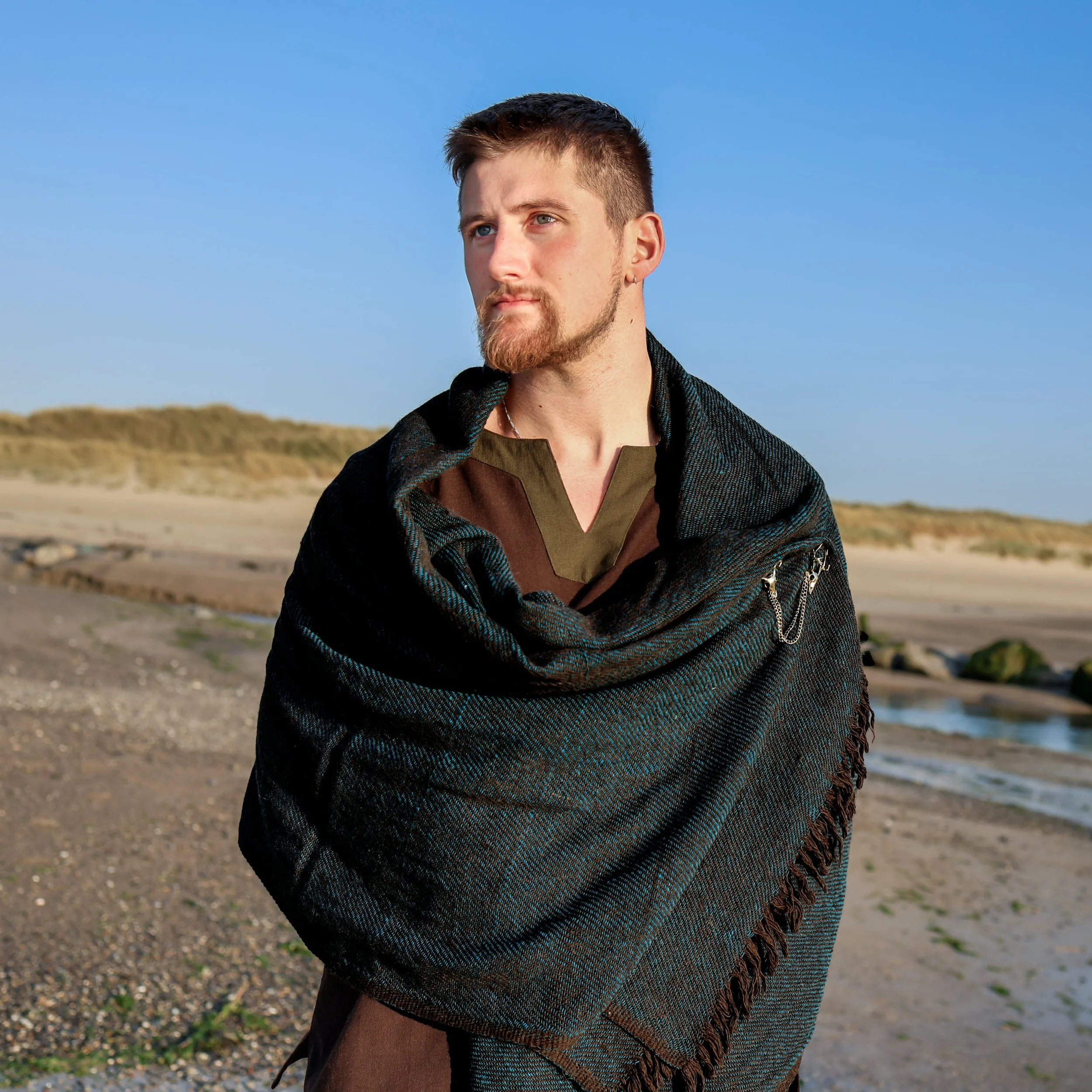 A man standing on a beach wearing a dark handwoven Celtic shawl with fringe detailing. The shawl is draped over his shoulders and fastened with a decorative brooch, evoking a Viking-inspired style.
