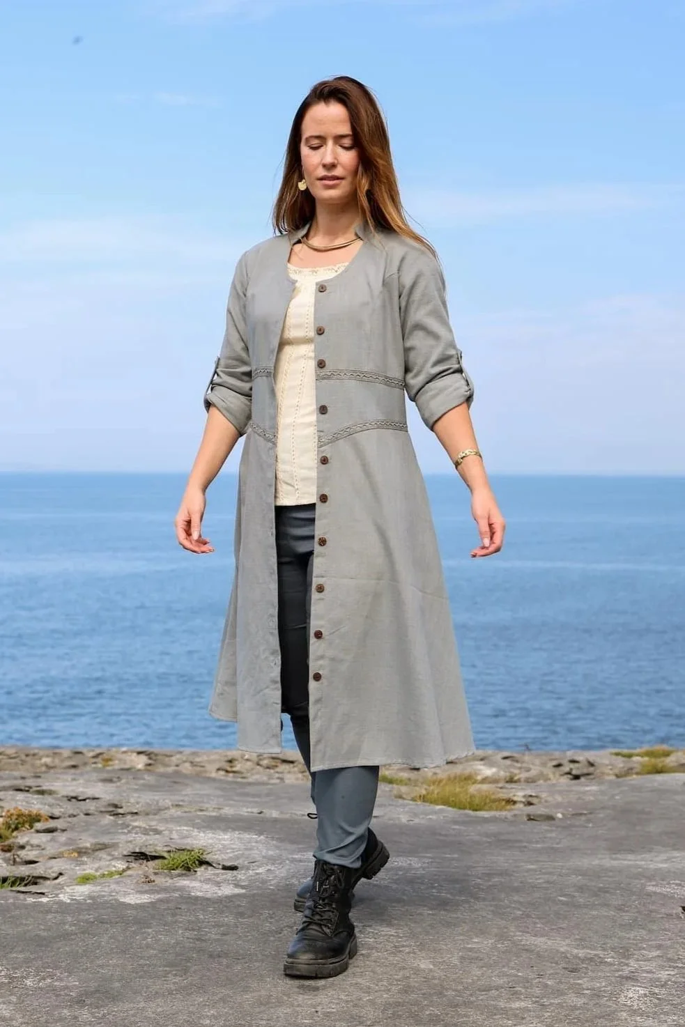 Model wearing a blue cotton button down dress with Celtic trim, styled as a midi dress with pockets and detailed button front dress closure.