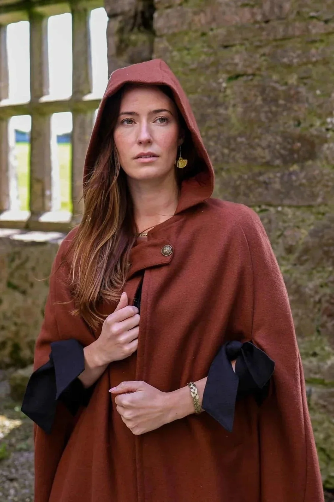 Woman wearing a hooded cape styled as a medieval cloak by Celtic Fusion, evoking a red riding hood silhouette in a rustic stone ruin setting