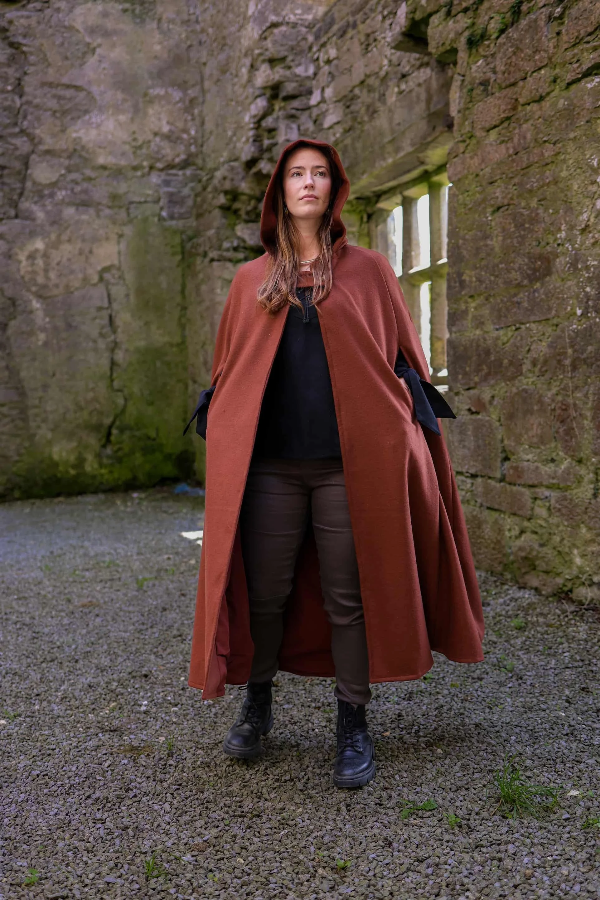 Woman wearing a hooded cape styled as a medieval cloak by Celtic Fusion, evoking a red riding hood silhouette in a rustic stone ruin setting