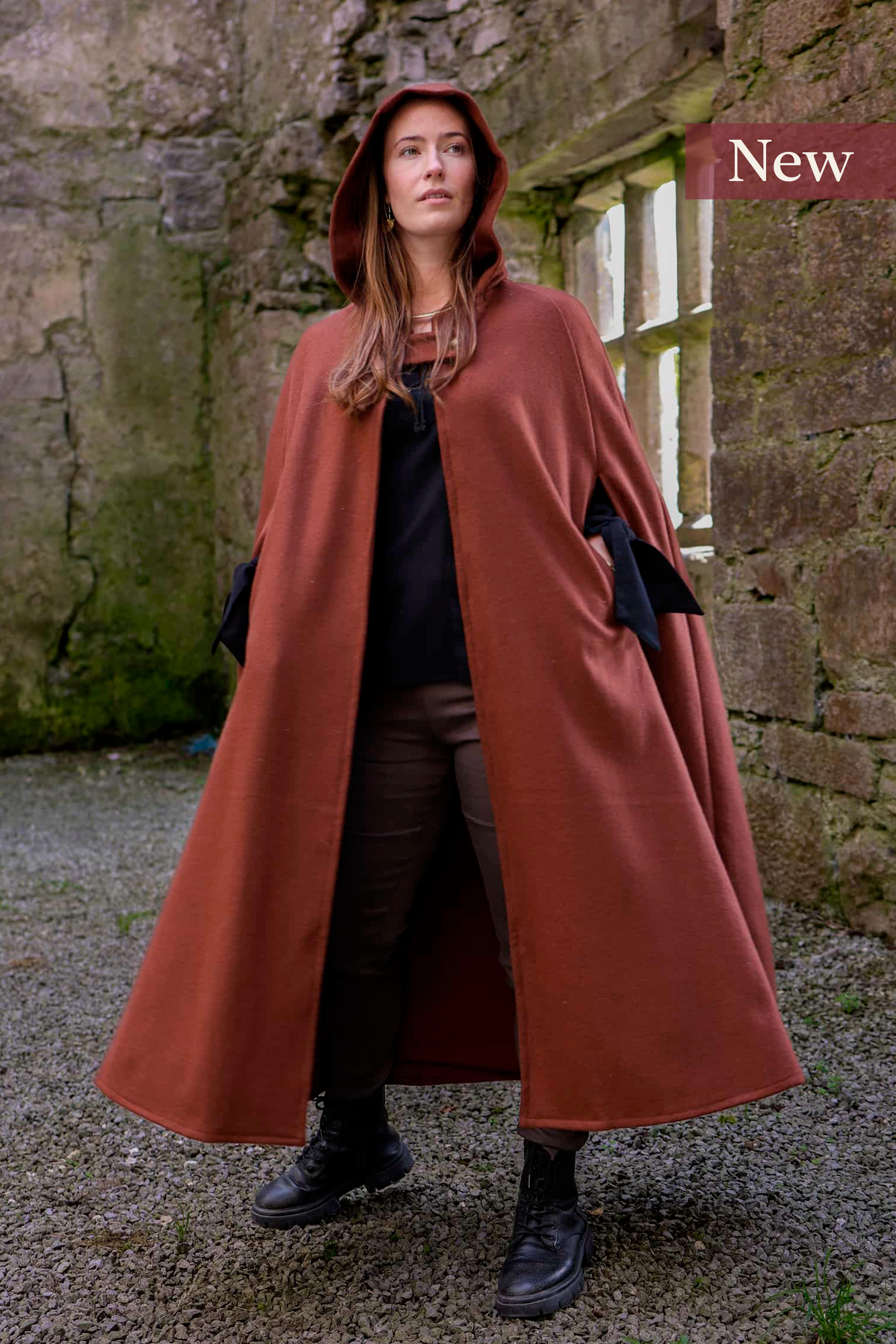 Woman wearing a hooded cape styled as a medieval cloak by Celtic Fusion, evoking a red riding hood silhouette in a rustic stone ruin setting