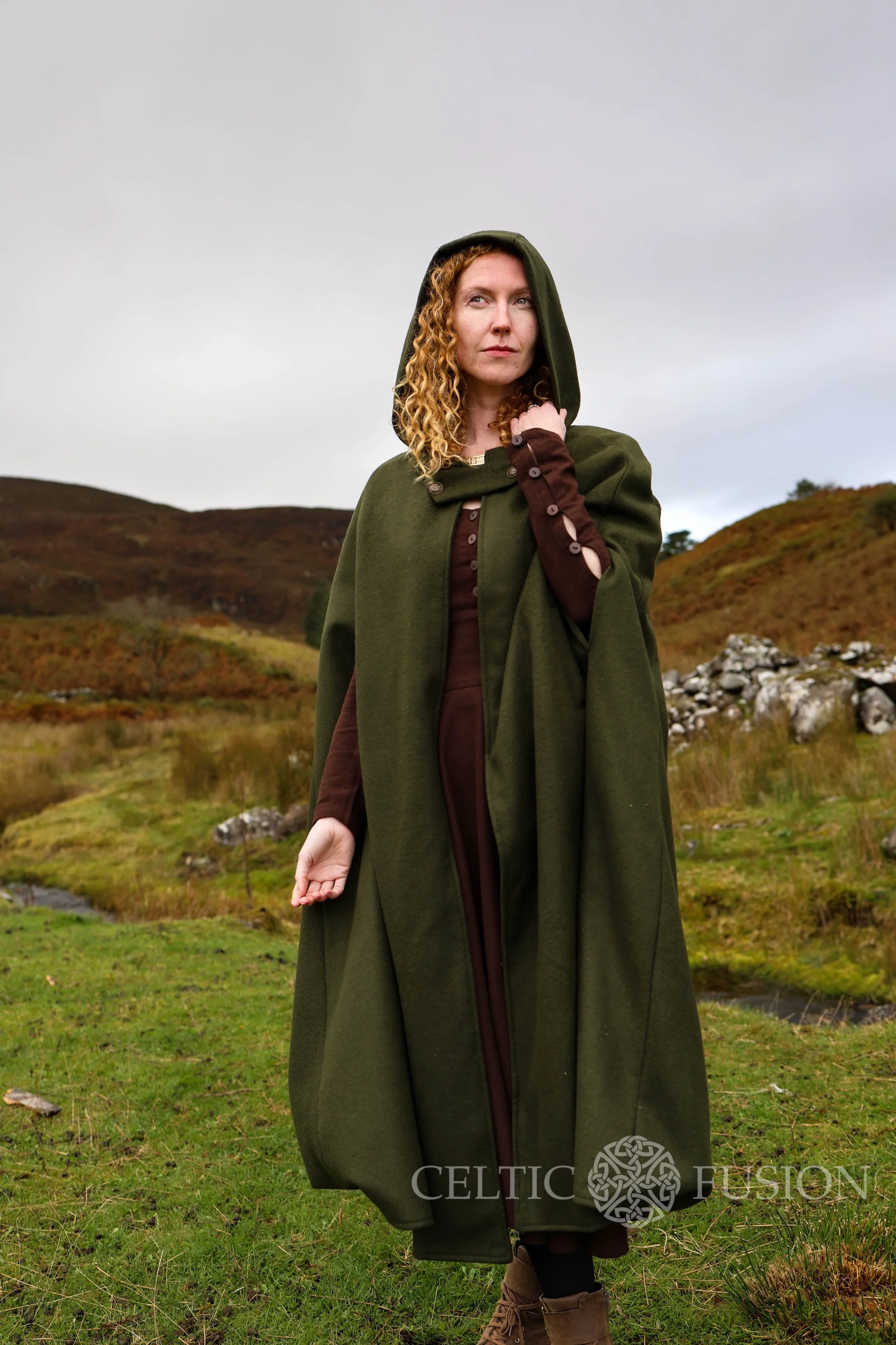 Woman wearing a hooded cape styled as a medieval cloak by Celtic Fusion, evoking a red riding hood silhouette in a rustic stone ruin setting