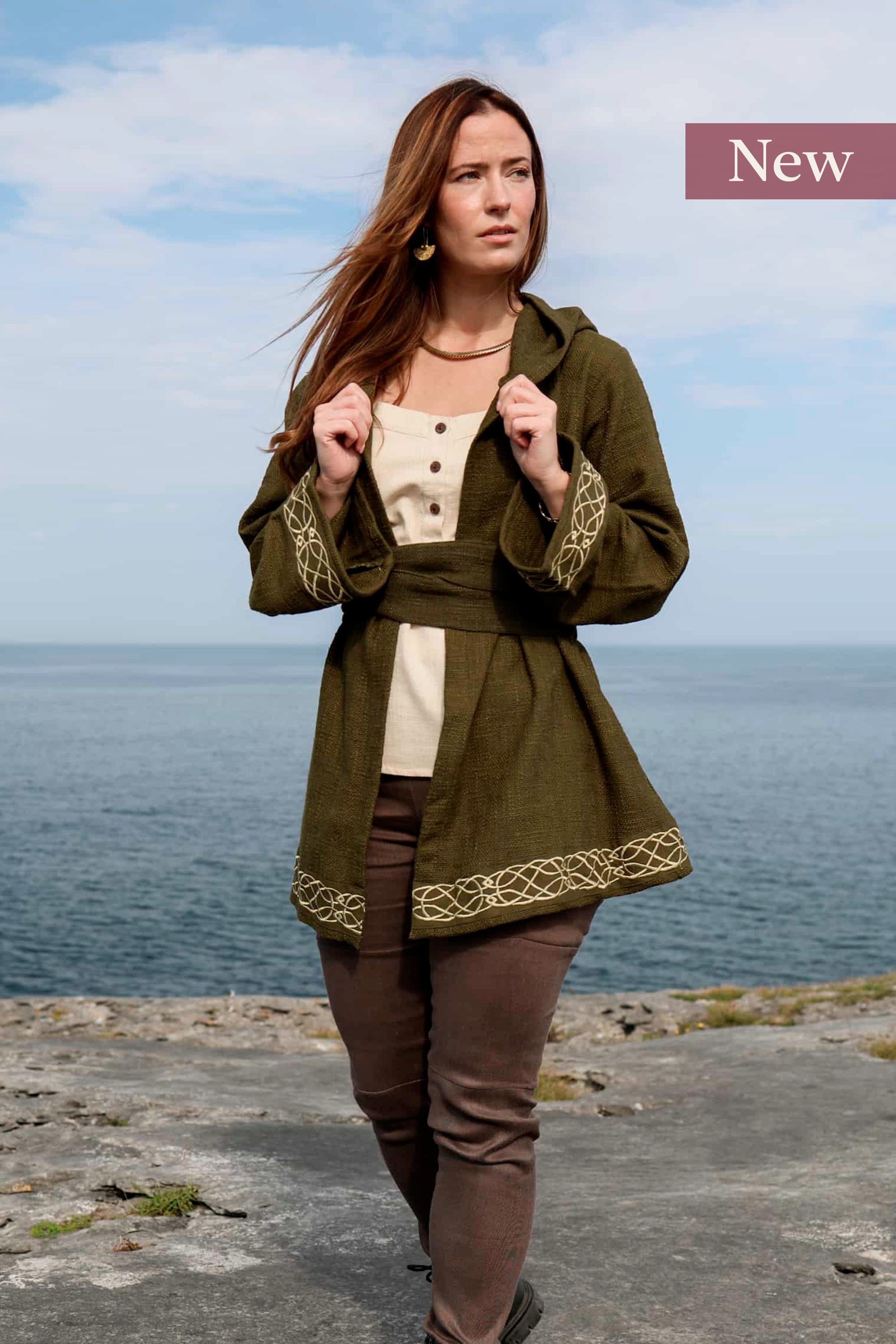 Woman wearing Celtic Fusions handwoven cotton hooded wrap cardigan with Celtic knotwork embroidery on cuffs and hem; belted at waist. Folklore cardigan photographed in a woodland setting.