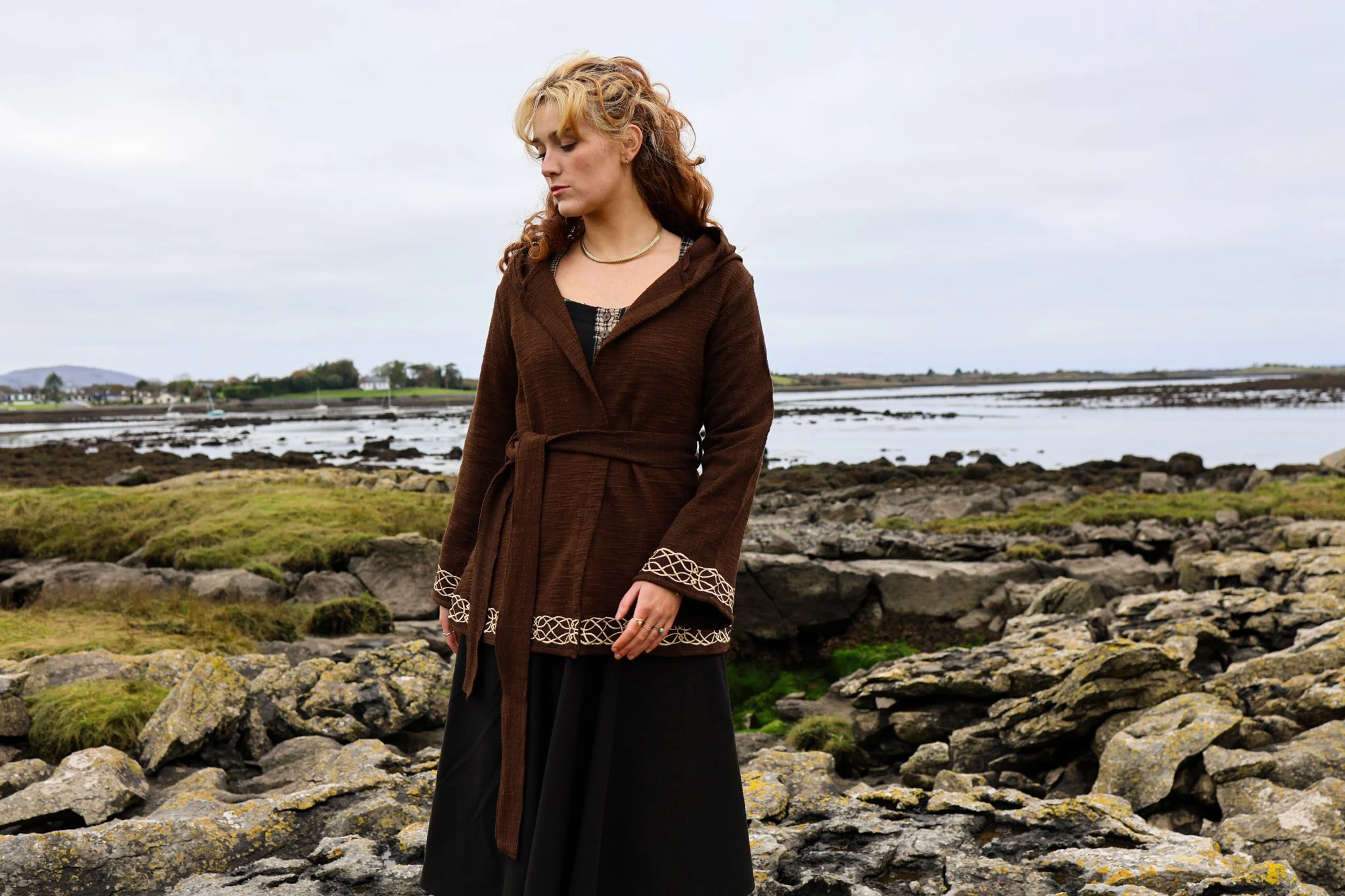 Woman wearing Celtic clothing handwoven wrap cardigan with Celtic knot embroidery on cuffs and hem; belted at waist. Folklore cardigan photographed in a woodland setting.