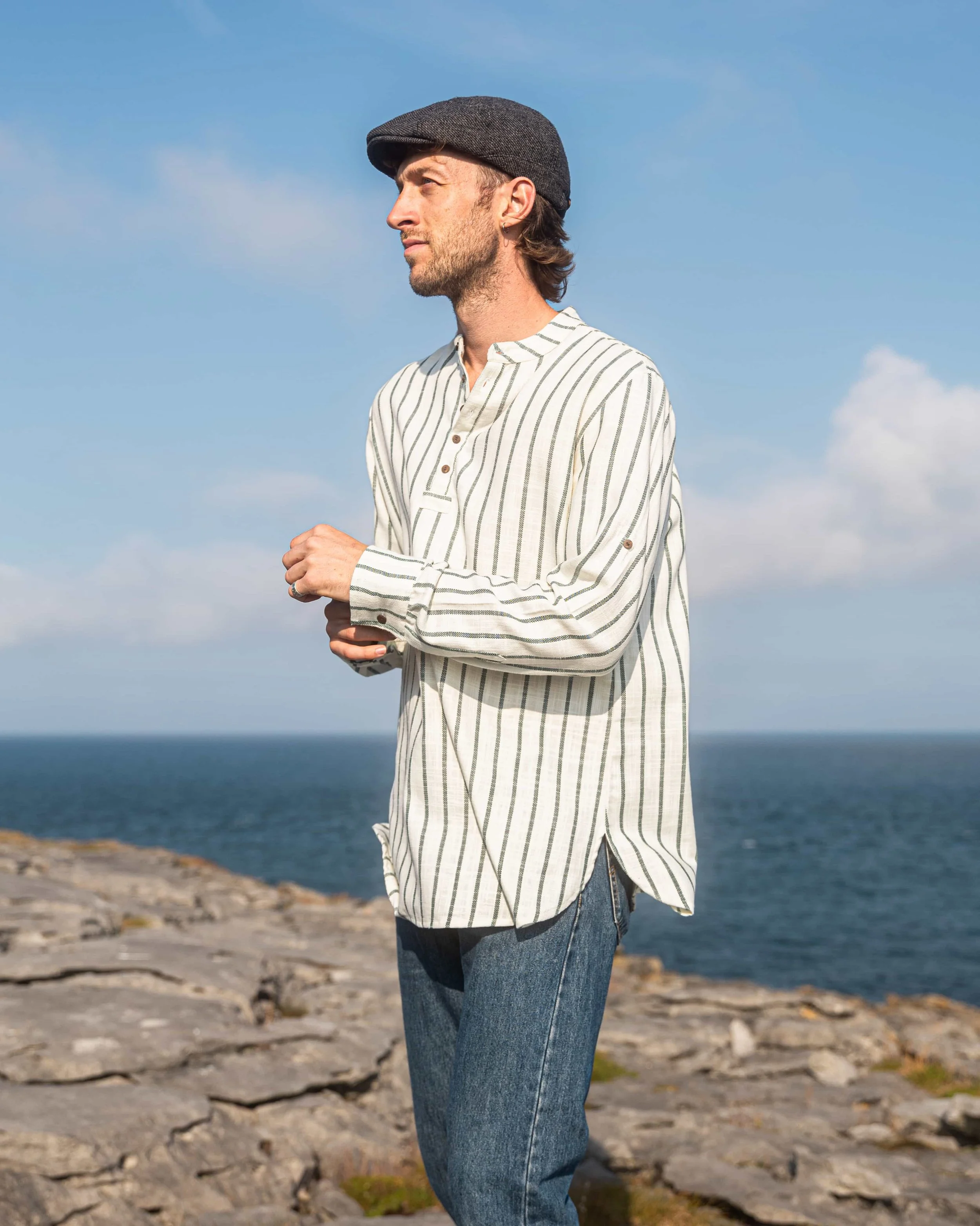 Model wearing a forest green and cream striped collarless shirt in 100% cotton, with coconut buttons and rolled sleeves, pictured by the Atlantic coast.