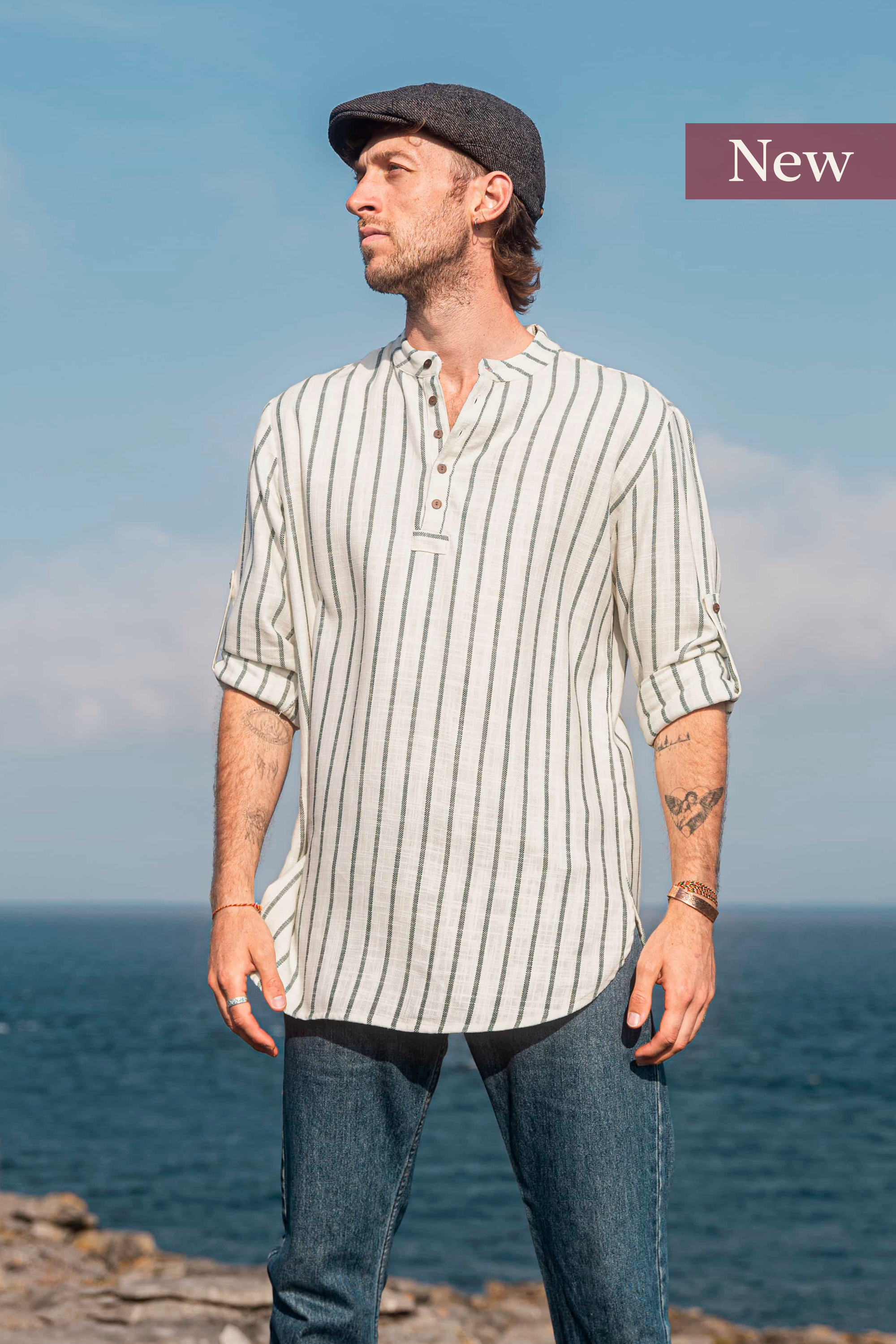 Model wearing a forest green and cream striped collarless shirt in 100% cotton, with coconut buttons and rolled sleeves, pictured by the Atlantic coast.