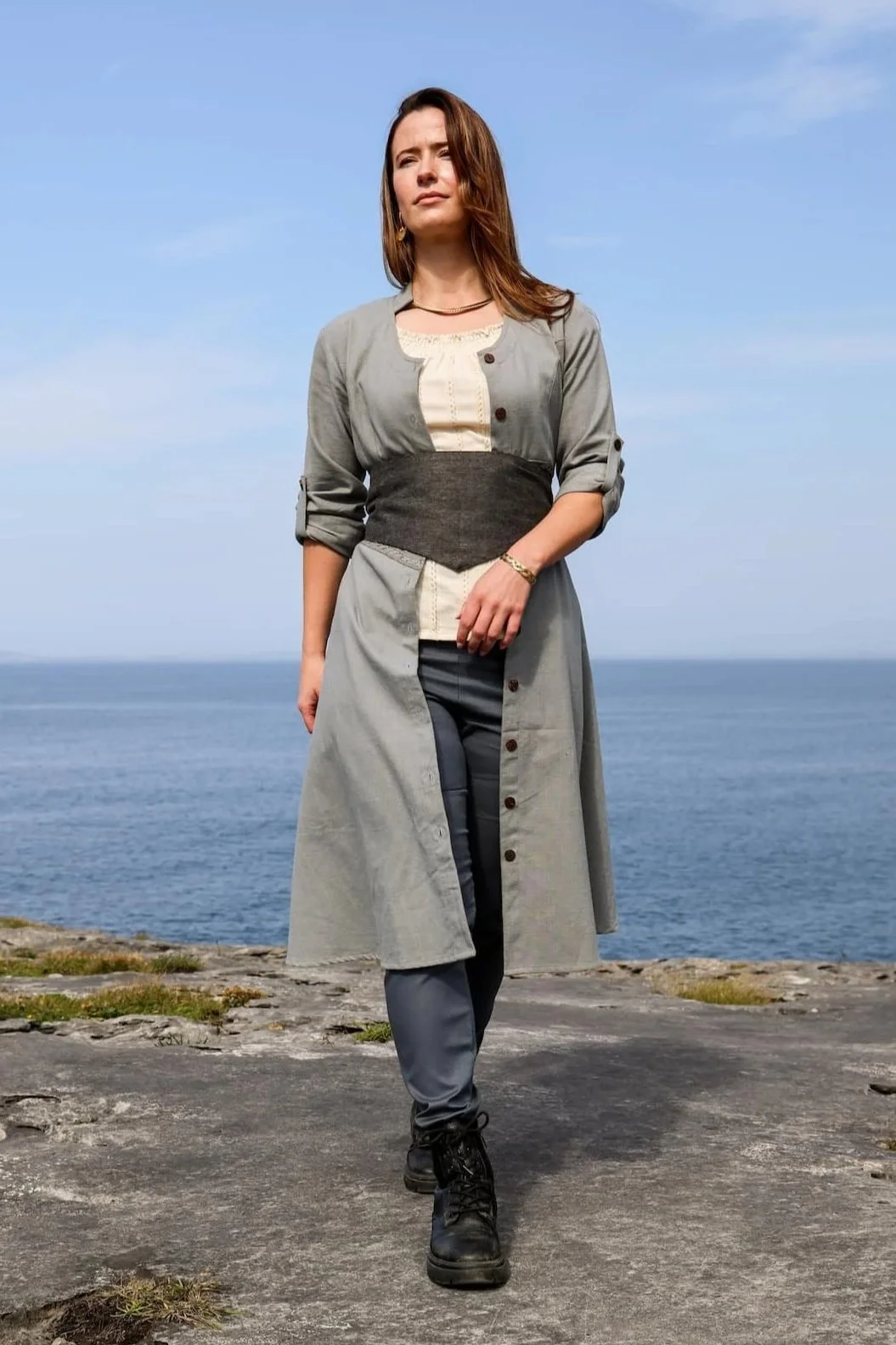 Model wearing a blue cotton button down dress with Celtic trim, styled as a midi dress with pockets and detailed button front dress closure.