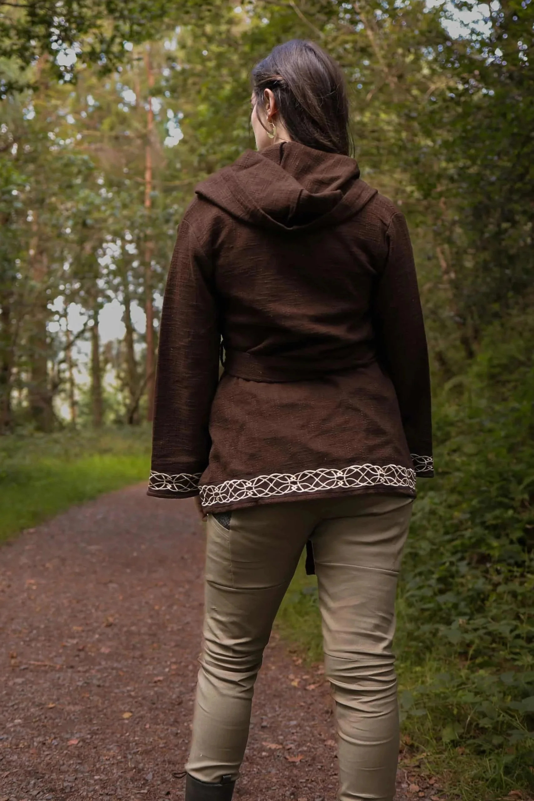 Woman wearing Celtic clothing handwoven wrap cardigan with Celtic knot embroidery on cuffs and hem; belted at waist. Folklore cardigan photographed in a woodland setting.