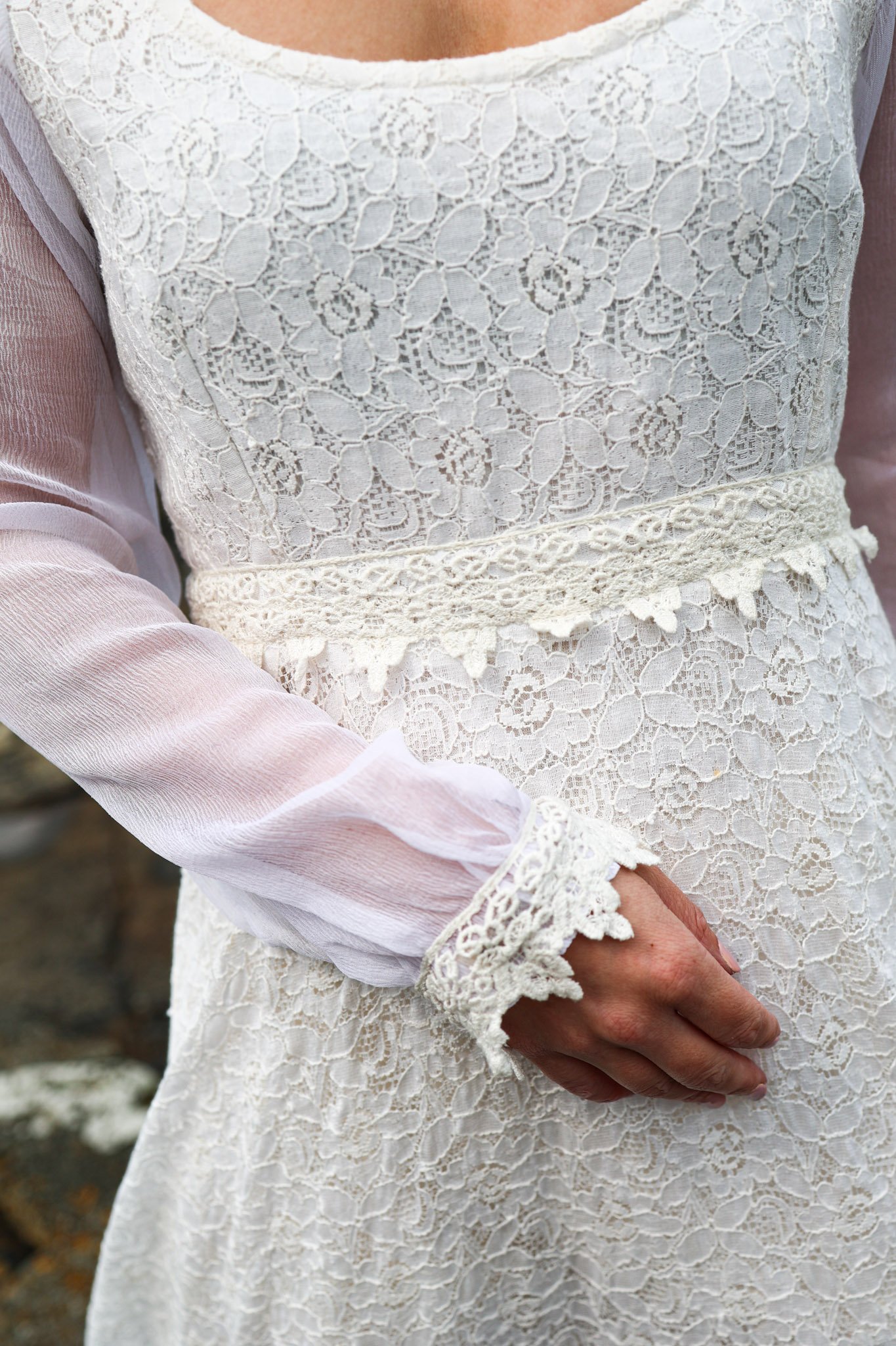 Annagh cotton lace wedding dress with bishop chiffon sleeves and a low scoop back, shown on a mannequin. Features delicate waist lace trim and a ruffled hem.