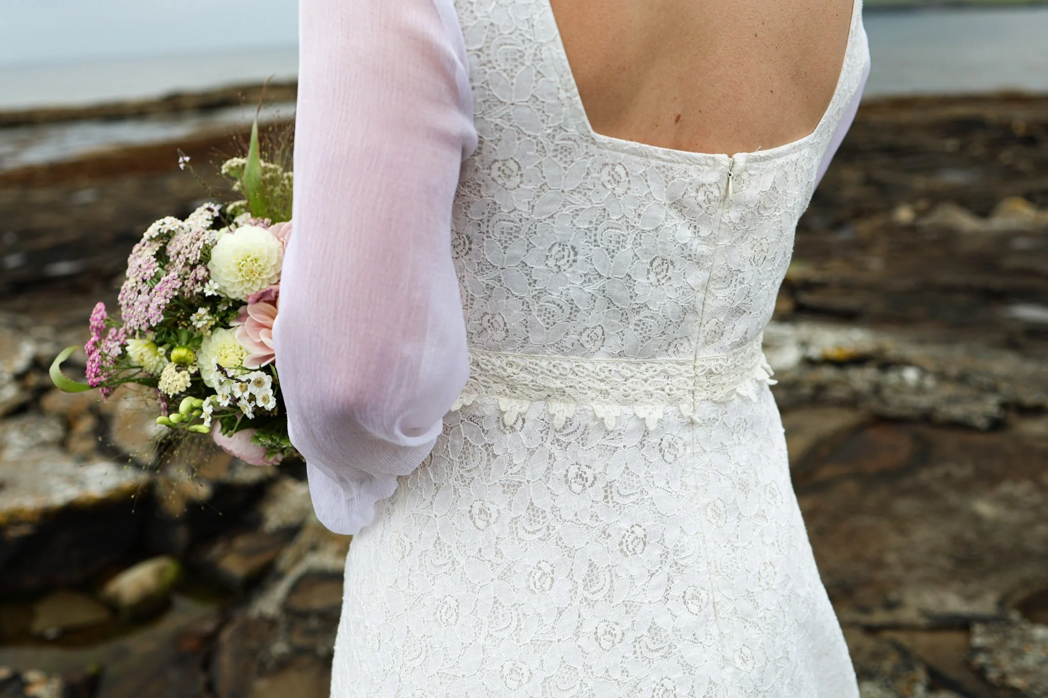 Annagh cotton lace wedding dress with bishop chiffon sleeves and a low scoop back, shown on a mannequin. Features delicate waist lace trim and a ruffled hem.